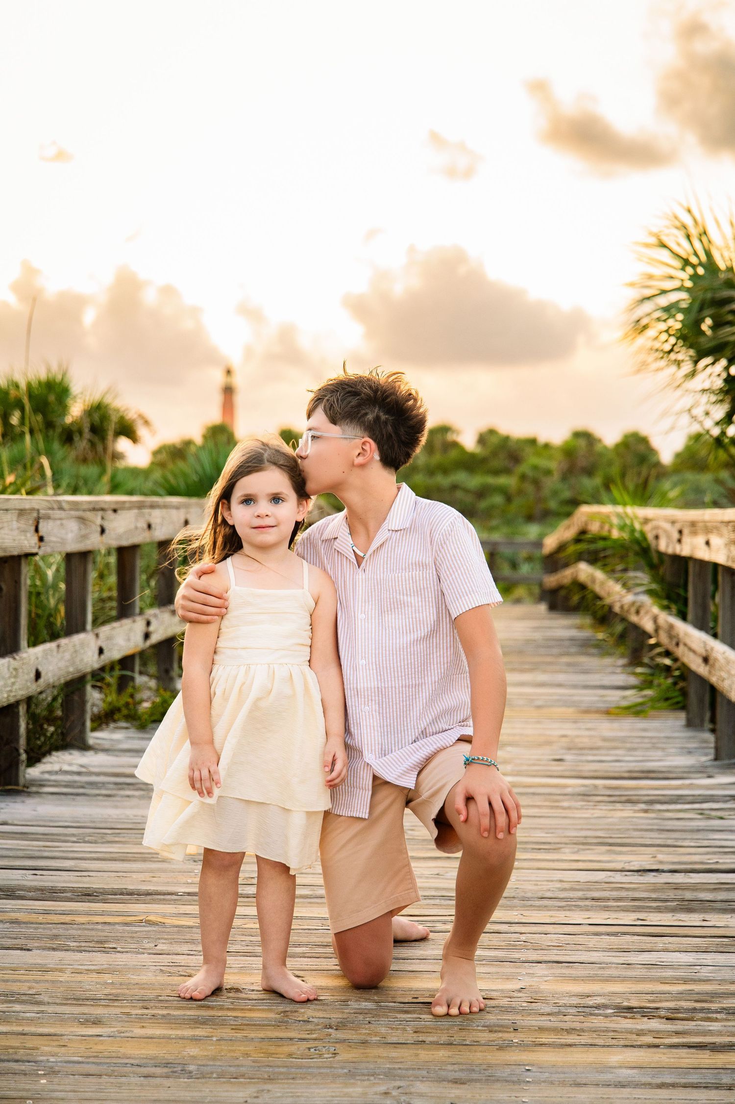 Older brother kneeling on a wooden boardwalk kissing younger sister on the cheek at sunset.