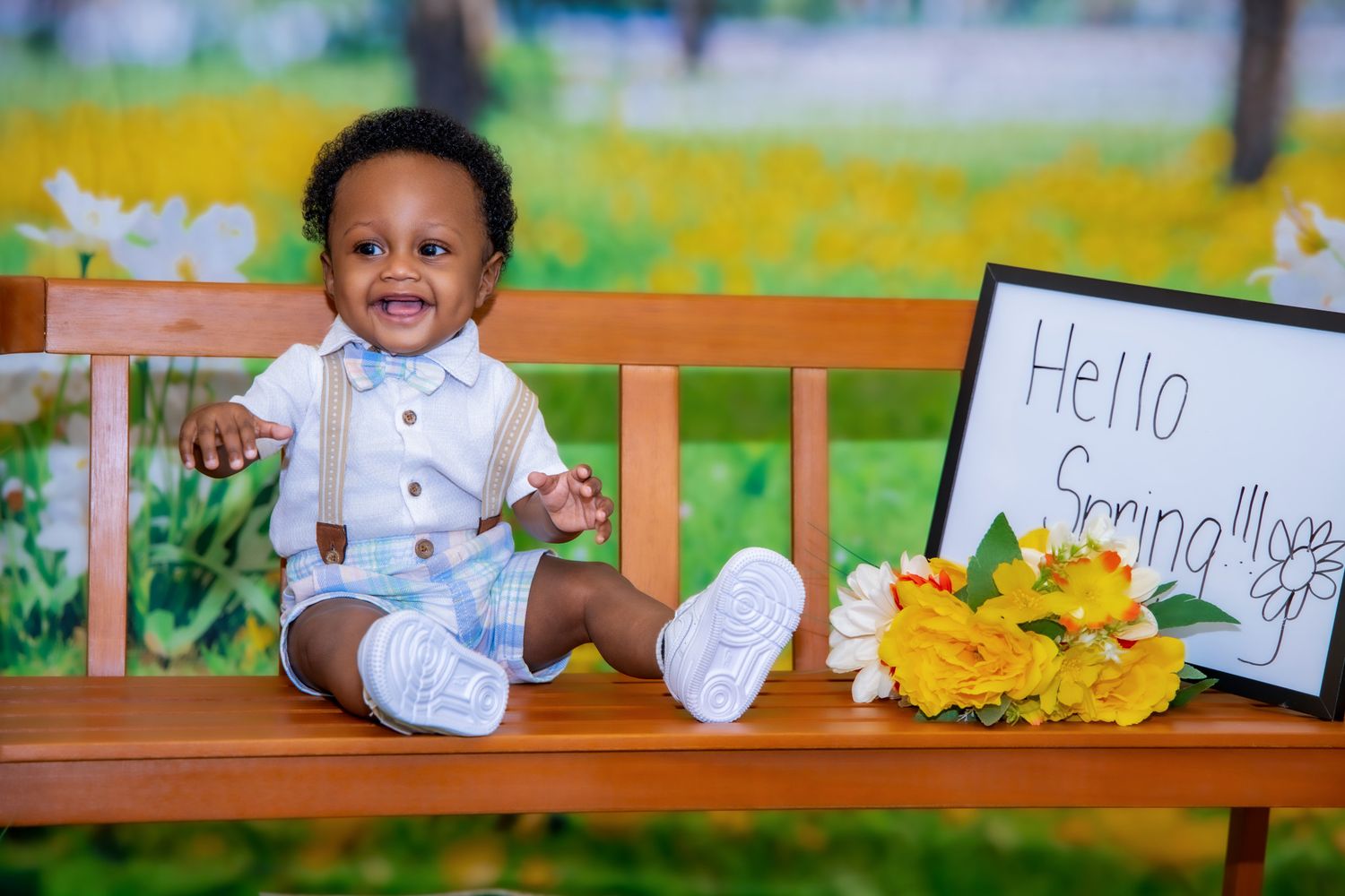 A baby wearing white sits on a wooden bench with yellow flowers and handwritten signs in an outdoor setting.