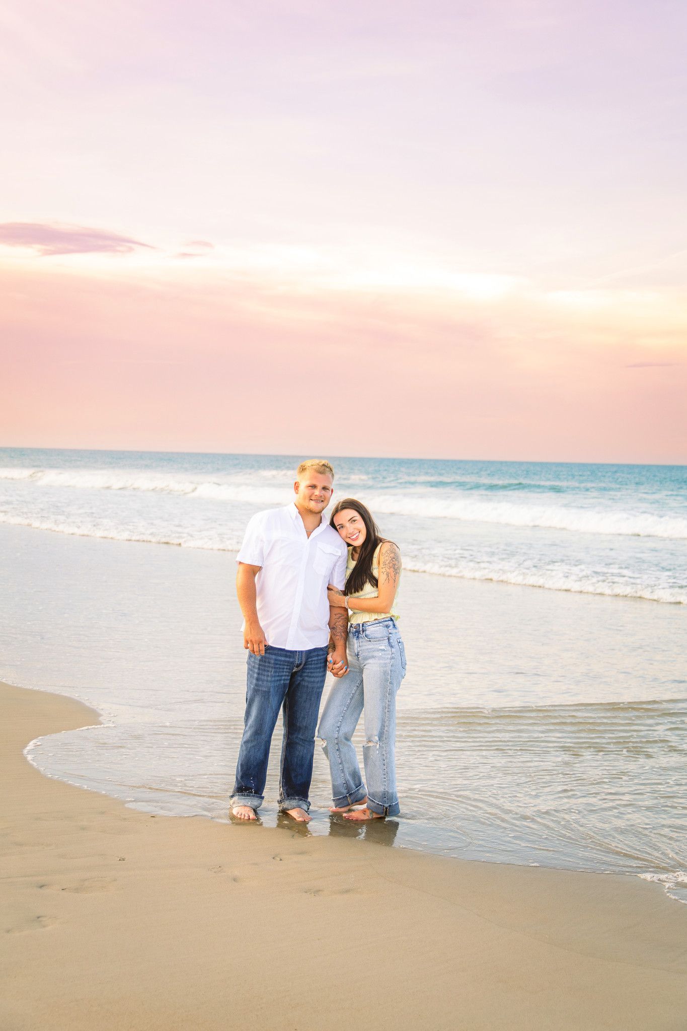 Couple holding hands on sandy beach at sunset with pink and purple sky and ocean waves behind them.