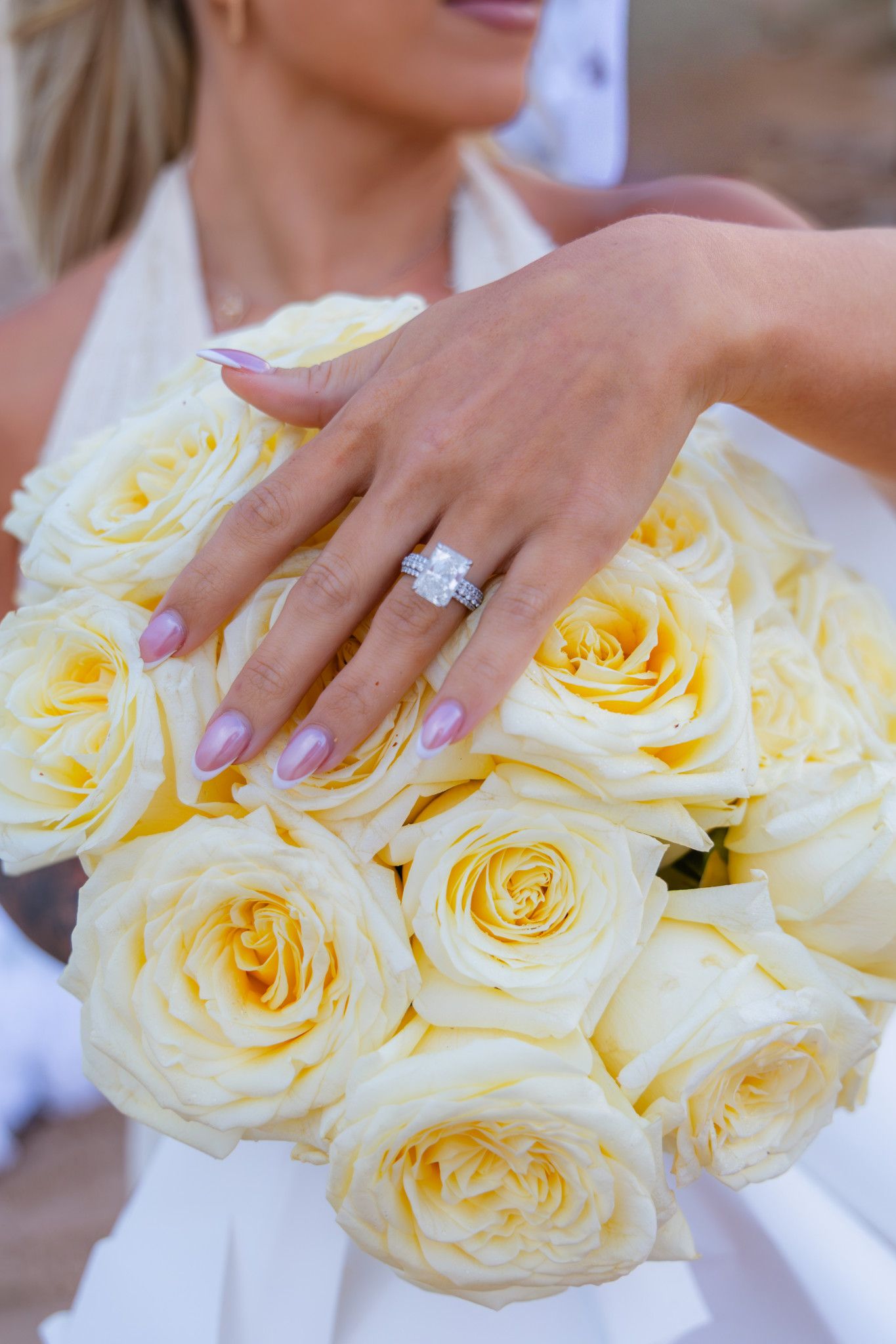 Bride in a white dress shows her square diamond engagement ring while holding a bouquet of pale yellow roses.
