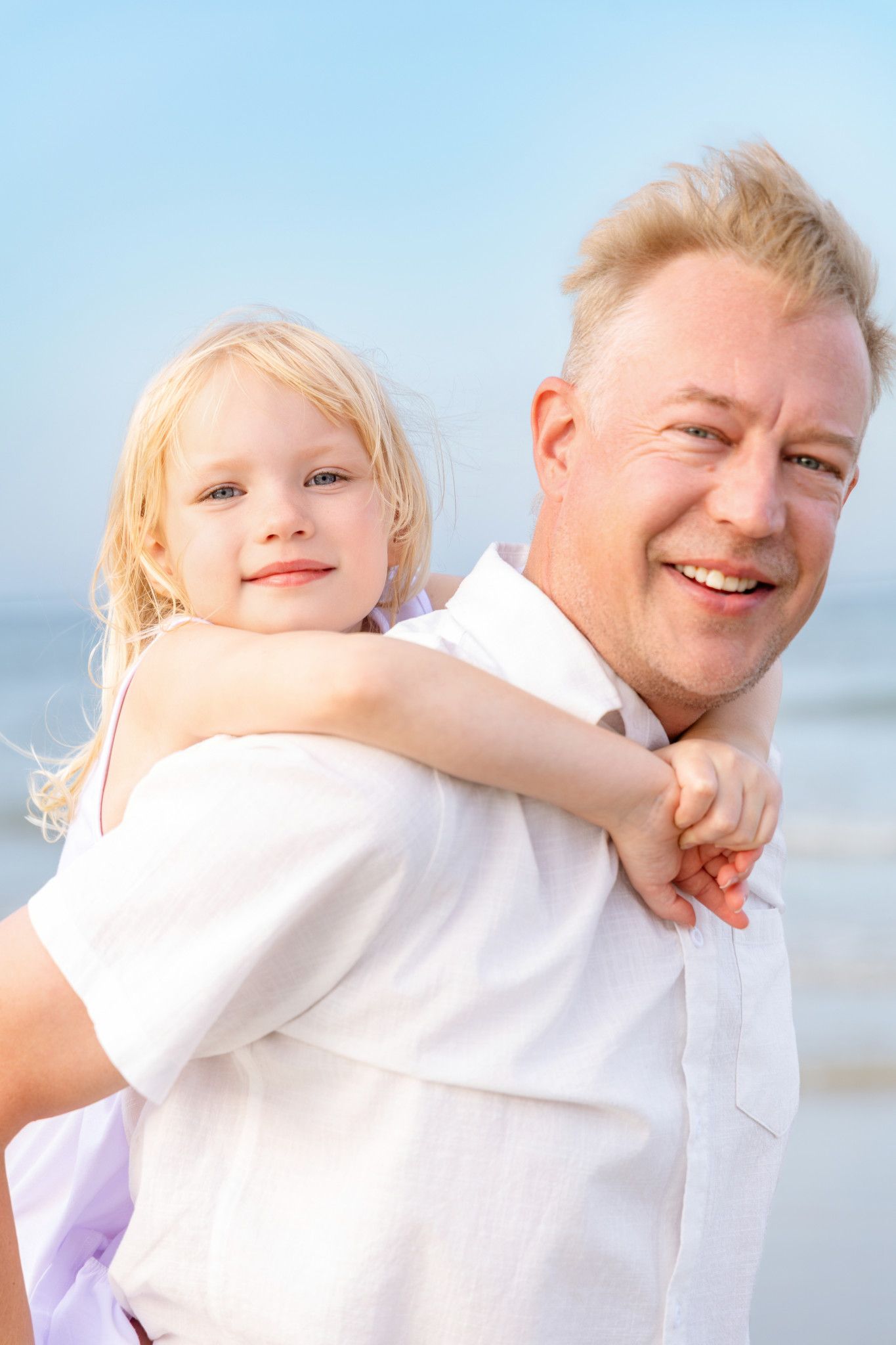 A smiling man giving a happy blonde toddler a piggyback ride on a sunny beach.