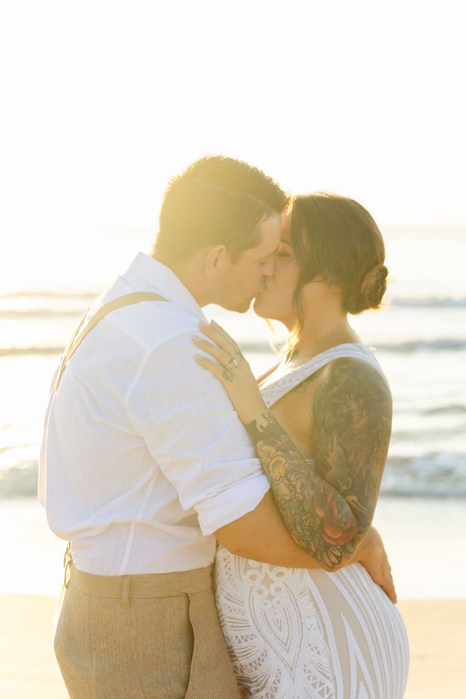 Tattooed bride and groom share a romantic kiss on a glowing sunlit beach.
