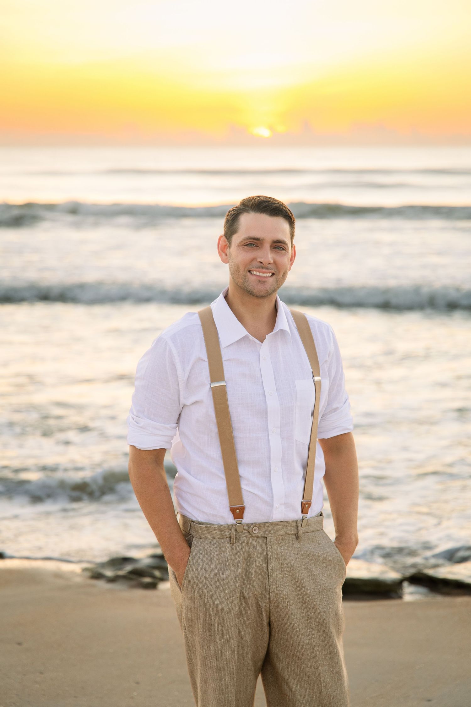 Smiling groom in white shirt and suspenders stands on beach during vibrant sunset.