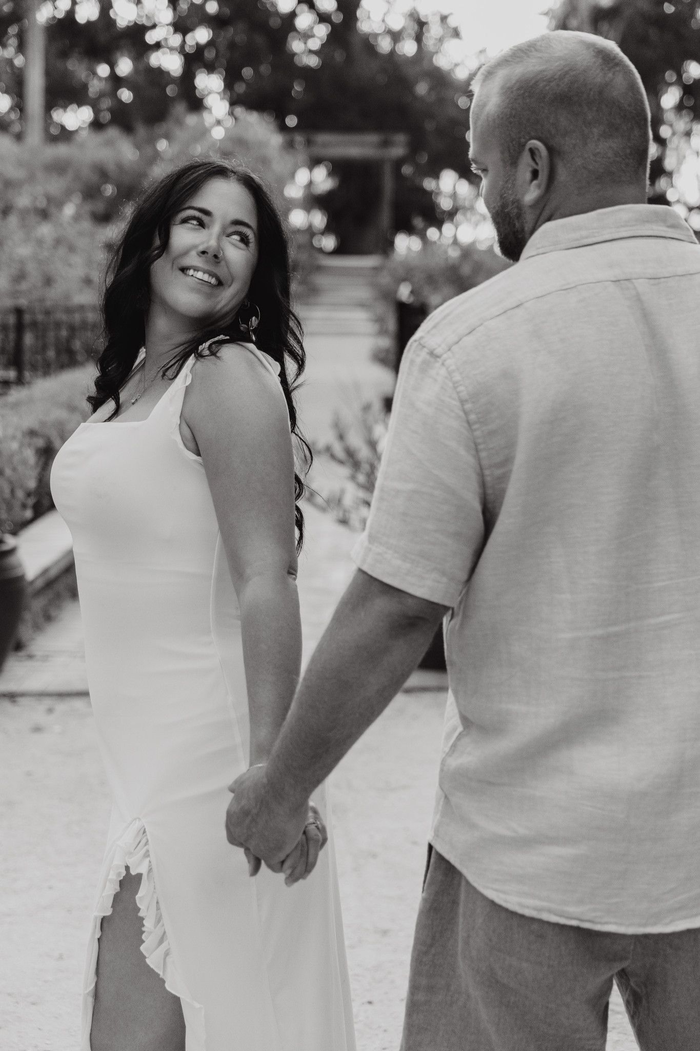 Black and white photo of a couple holding hands, woman in white dress smiling at man in casual shirt outdoors.