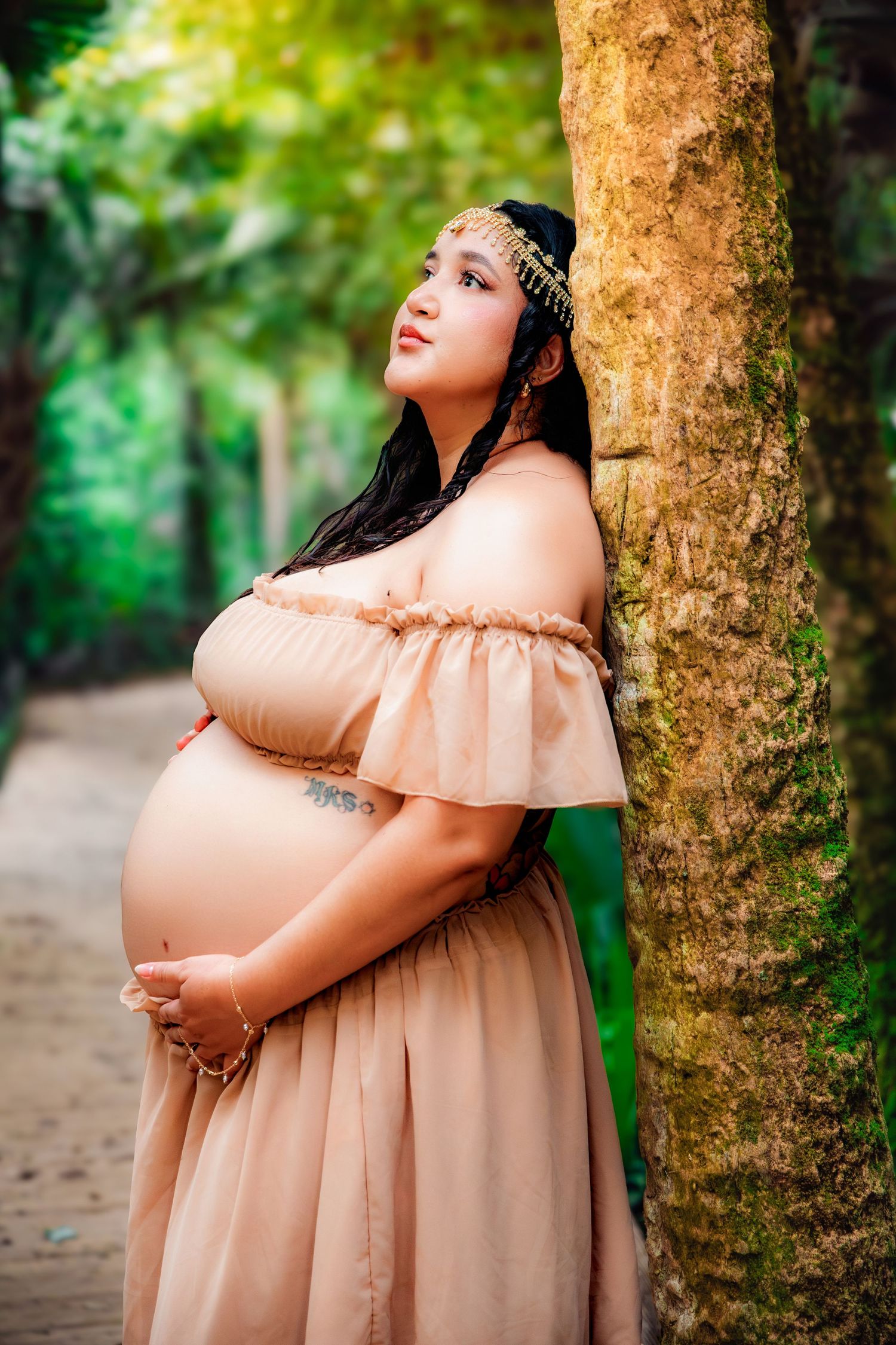 Pregnant woman in peach off-shoulder gown leaning against tree in lush green park, wearing gold headpiece.