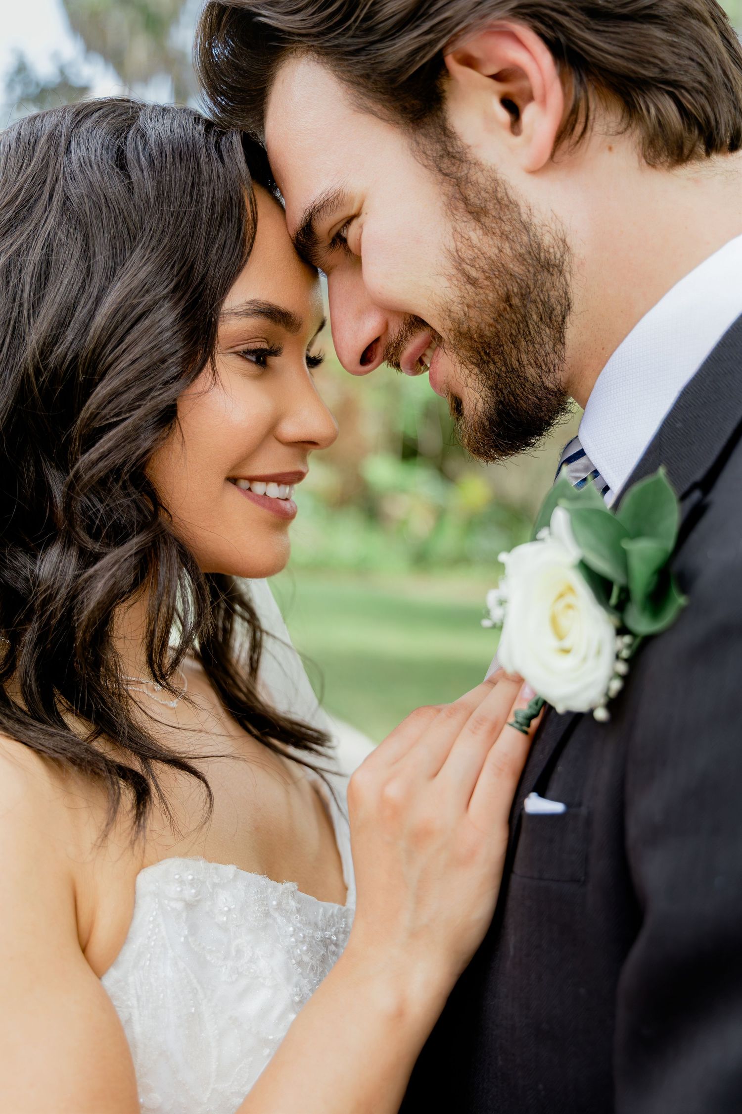 A bride and groom share an intimate moment, touching foreheads, as she holds his white rose boutonniere on their wedding day.