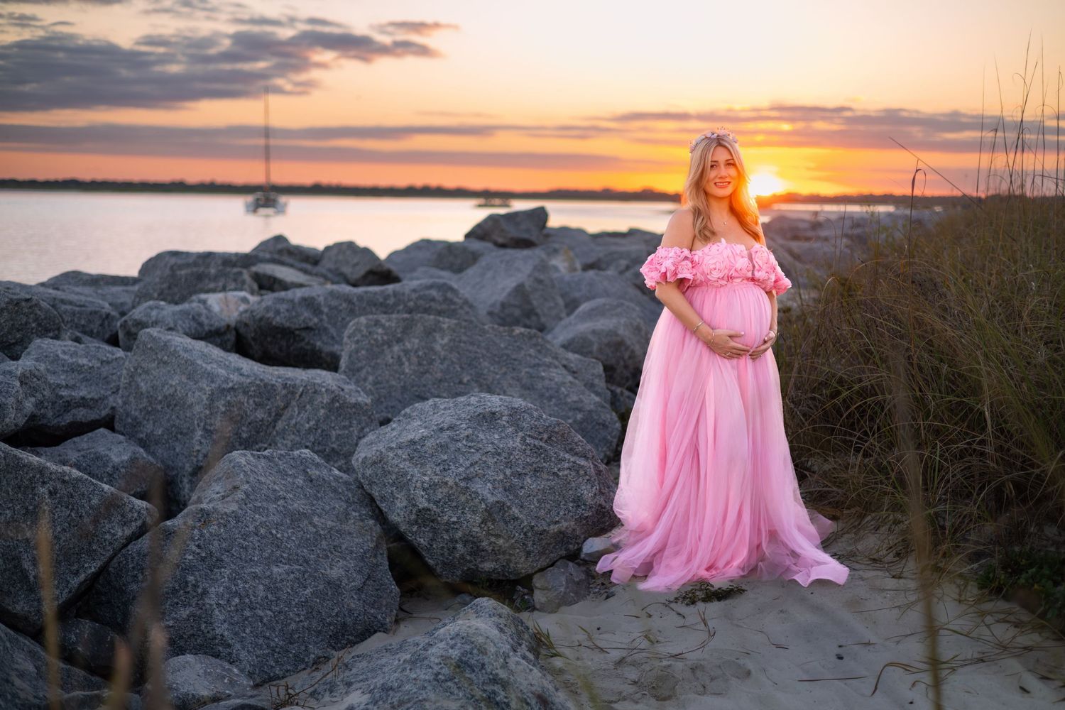 A figure in a flowing pink dress stands among coastal rocks during a dramatic sunset over the ocean.