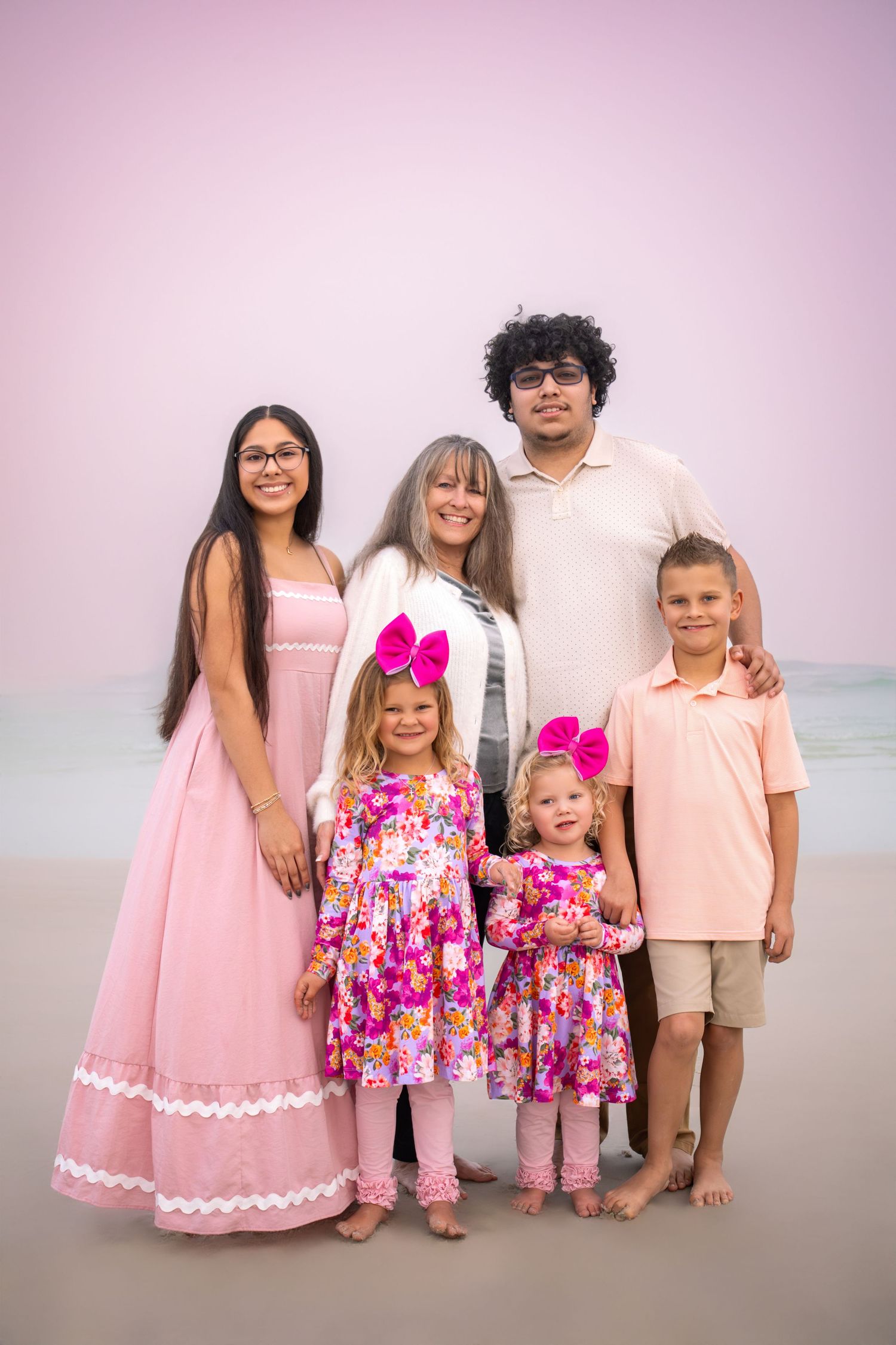 A family group in coordinating pink and floral outfits poses together on a beach at sunset.