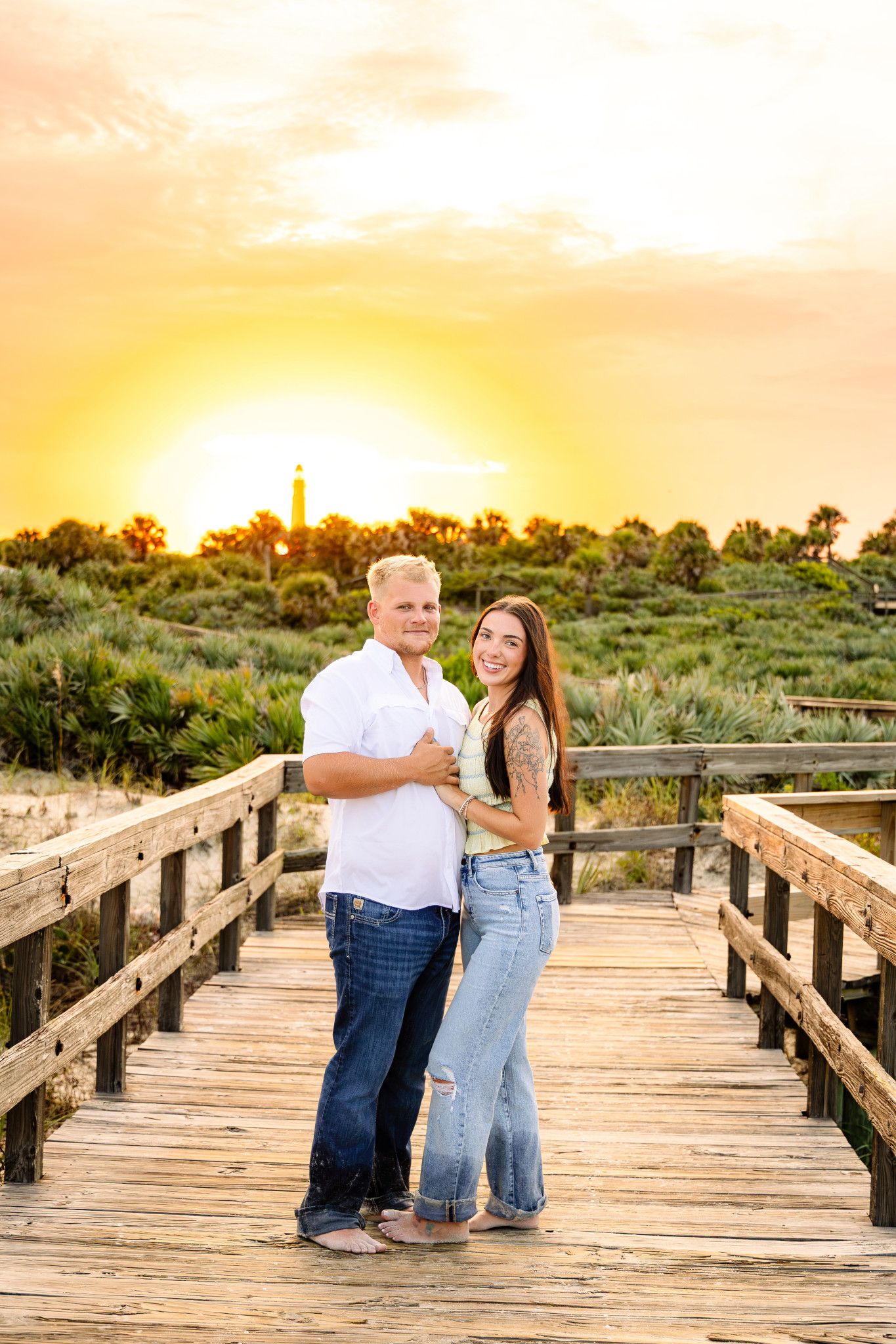 Couple embracing on a wooden boardwalk at golden sunset with a lighthouse and coastal dunes in the background.