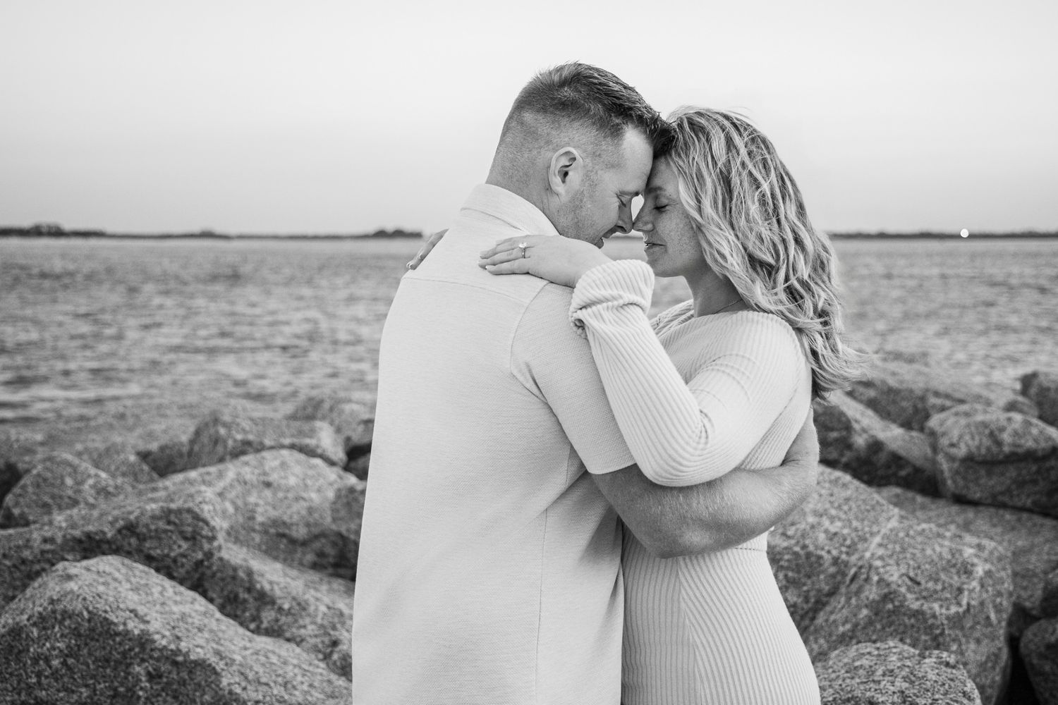 A romantic black and white photo series of a couple embracing on a rocky beach at sunset.