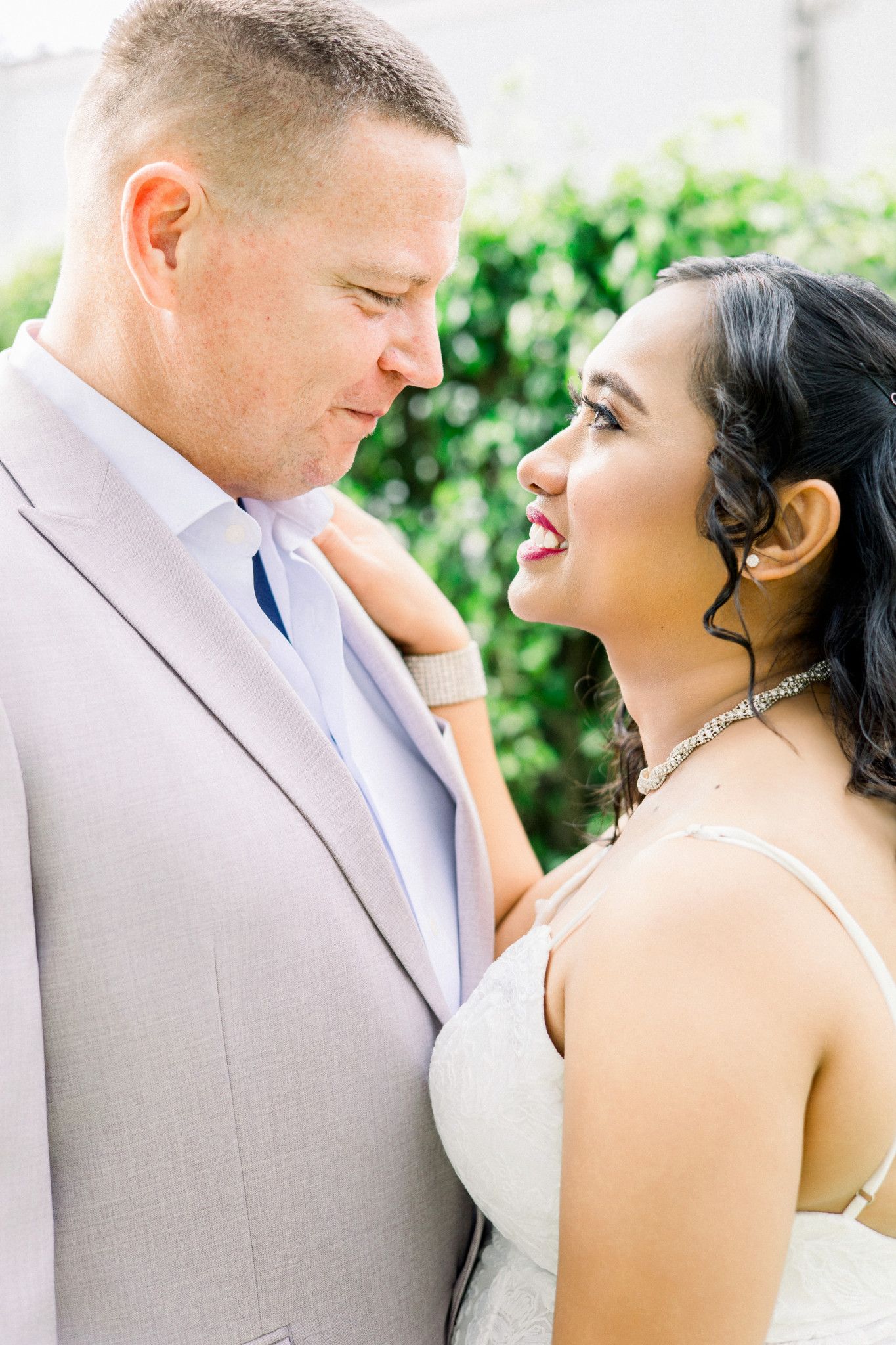 A bride and groom share an intimate moment, smiling at each other, dressed in wedding attire outdoors.