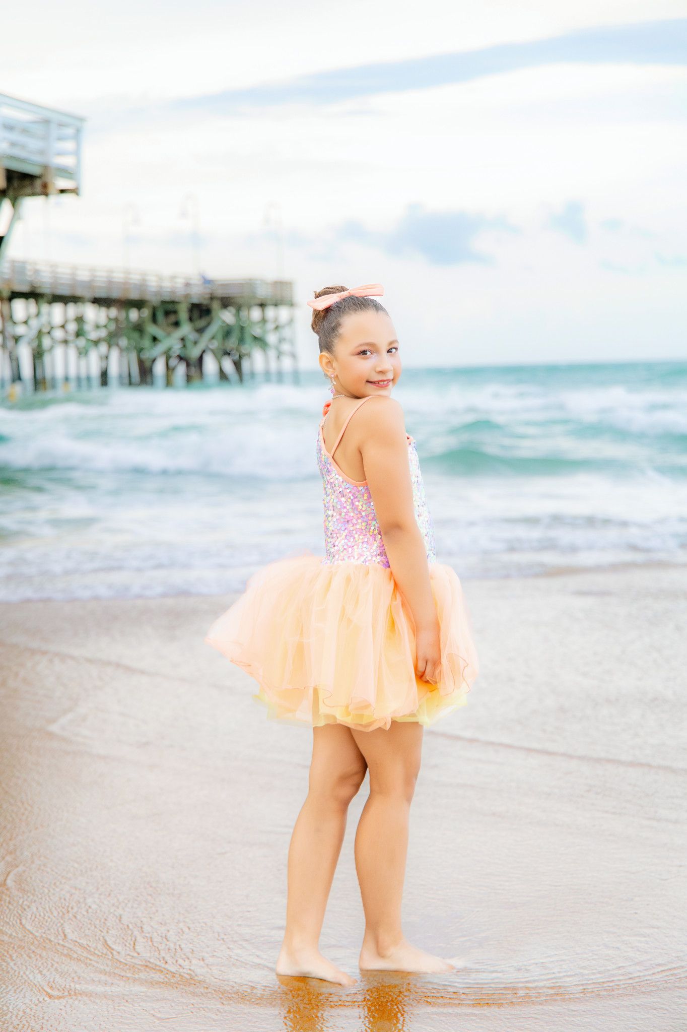 Young girl in pink sequin top and tulle skirt stands barefoot on sandy beach near pier with ocean waves.