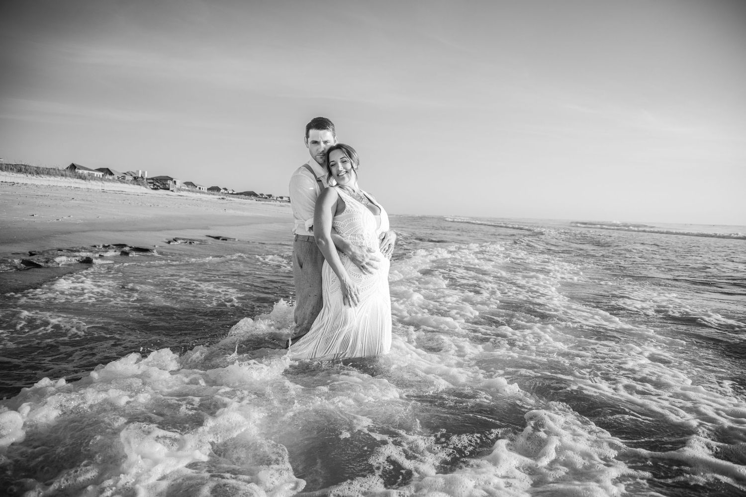 Black and white photo of a couple embracing in ocean waves on a beach, woman in flowing white dress.