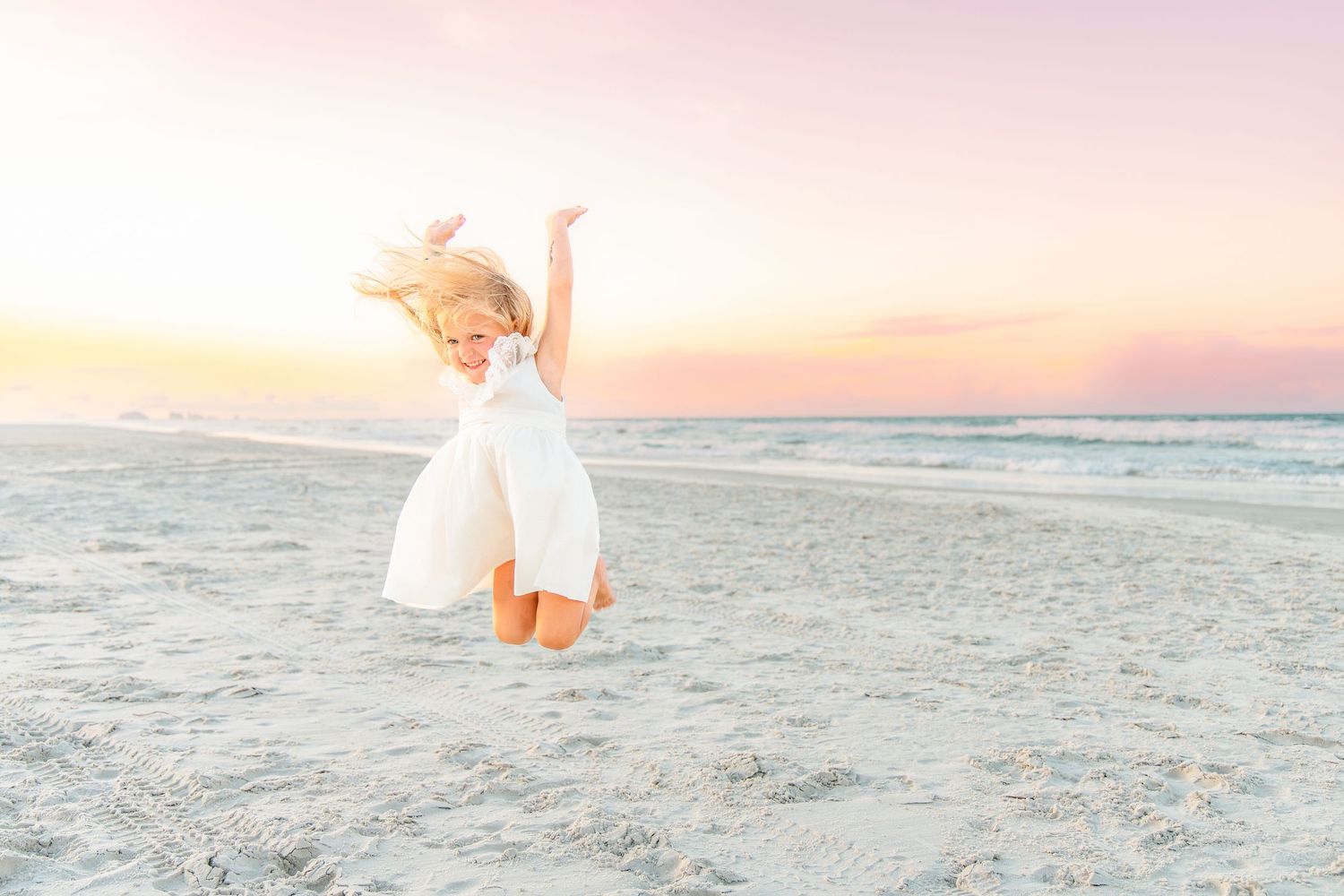 A joyful young girl in a white dress jumps on a sandy beach at sunset with arms raised.