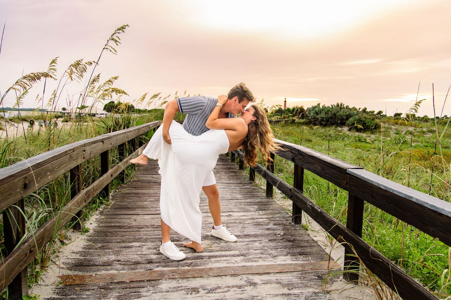 Couple sharing a romantic dip kiss on a wooden boardwalk surrounded by sea oats at sunset.