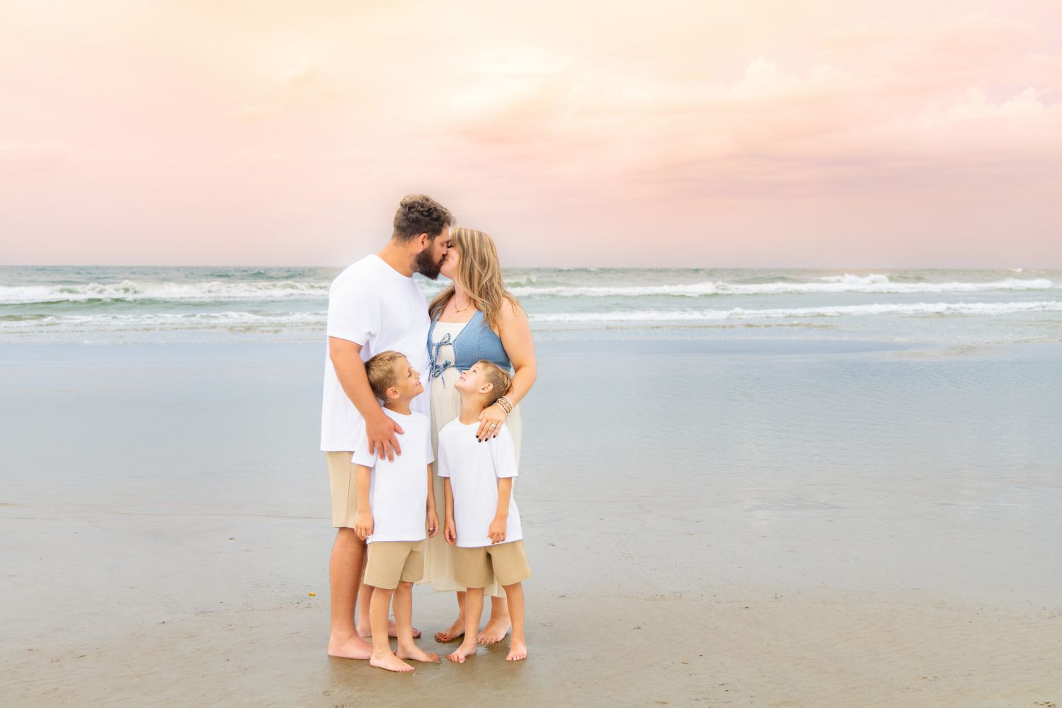 Family of four sharing a tender moment on a sandy beach at sunset with soft pink sky.