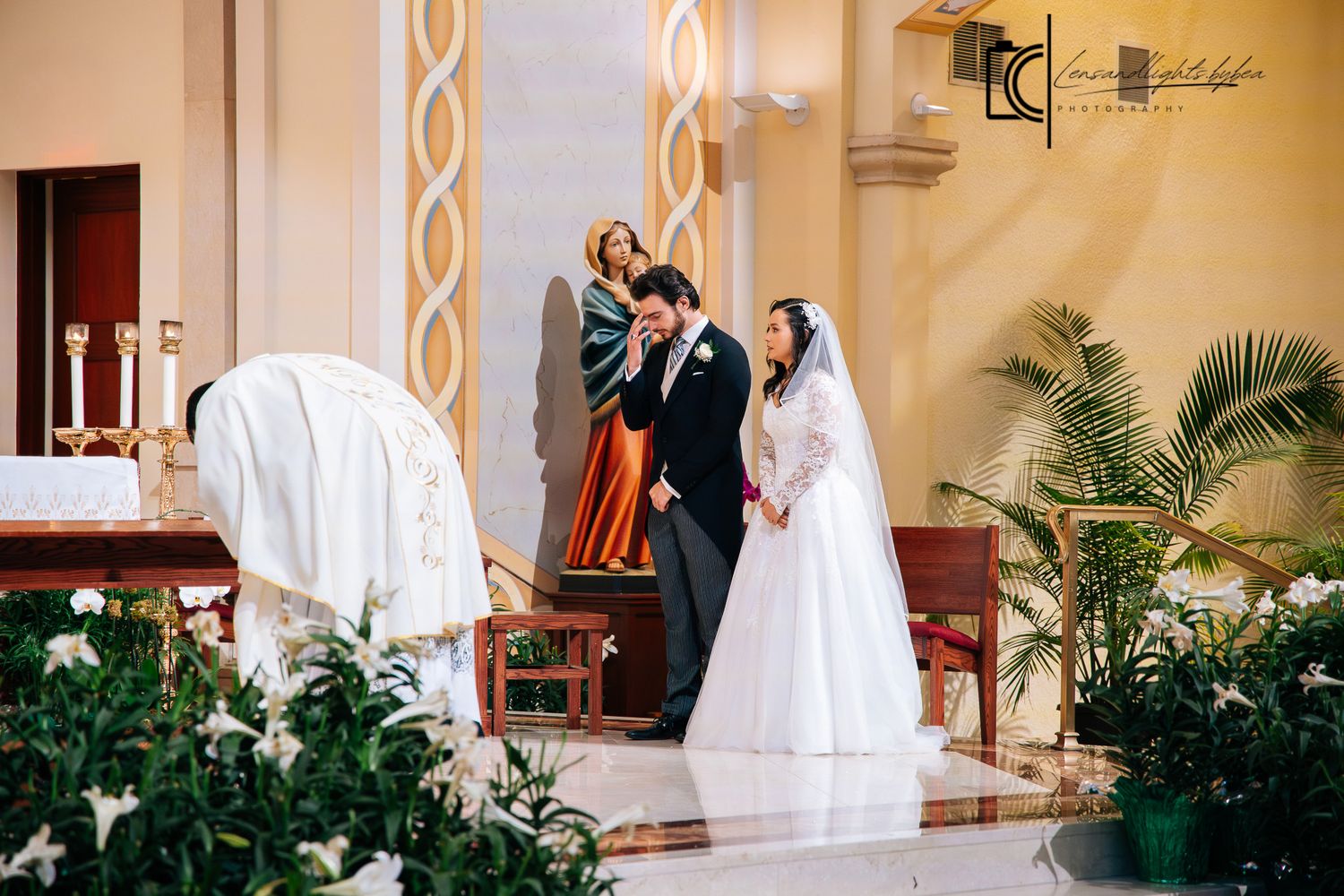 Bride and groom stand before a priest during a Catholic wedding ceremony in an elegant church.