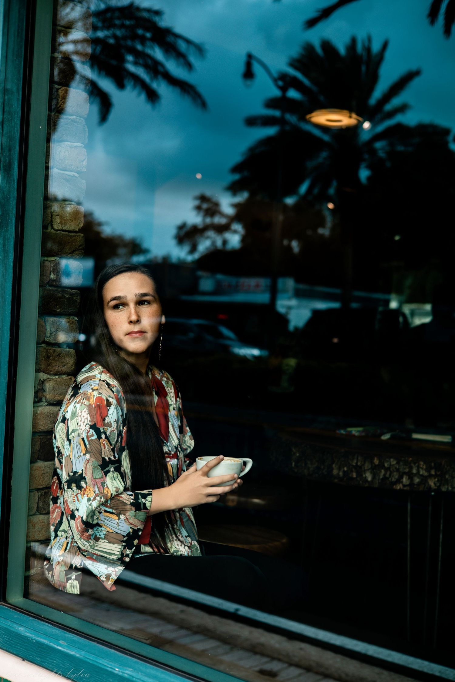 Person in floral print dress gazing through window with palm trees reflected.