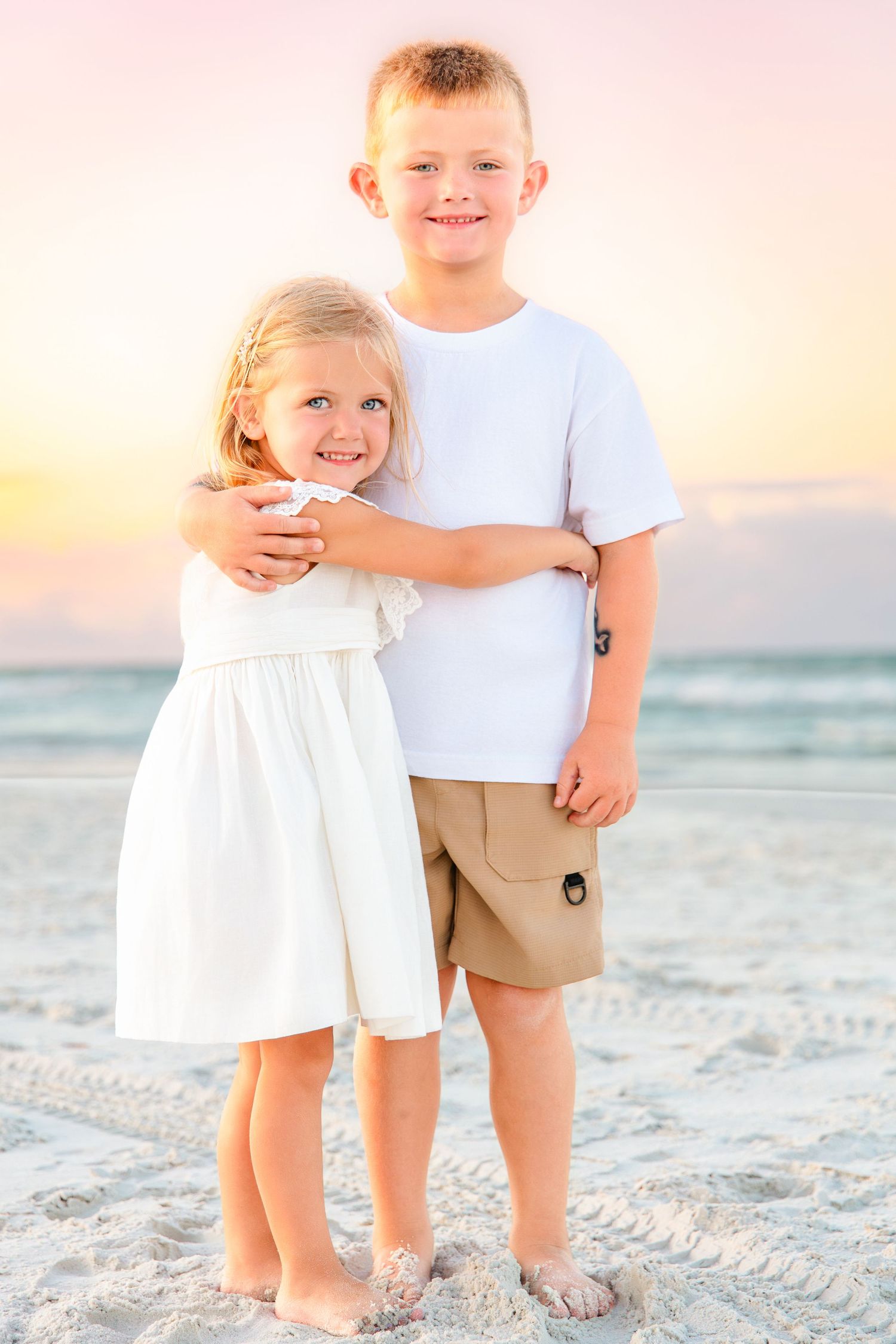 Two young siblings smile together on a sandy beach at sunset, boy in white shirt hugging girl in white dress.