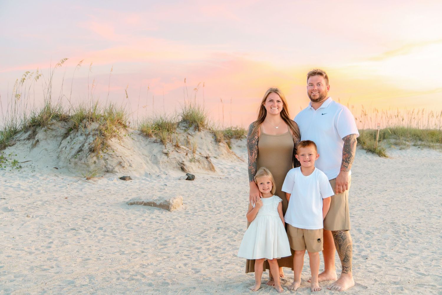 Family of four posing on a sandy beach at sunset with sea oats and pastel sky in the background.