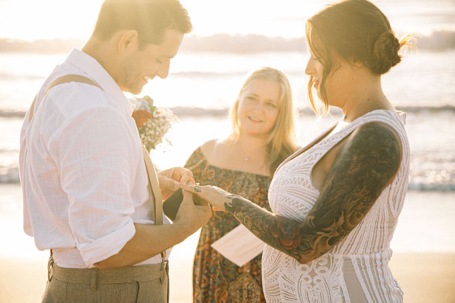 Tattooed bride and groom exchanging rings during outdoor beach wedding ceremony at sunset.