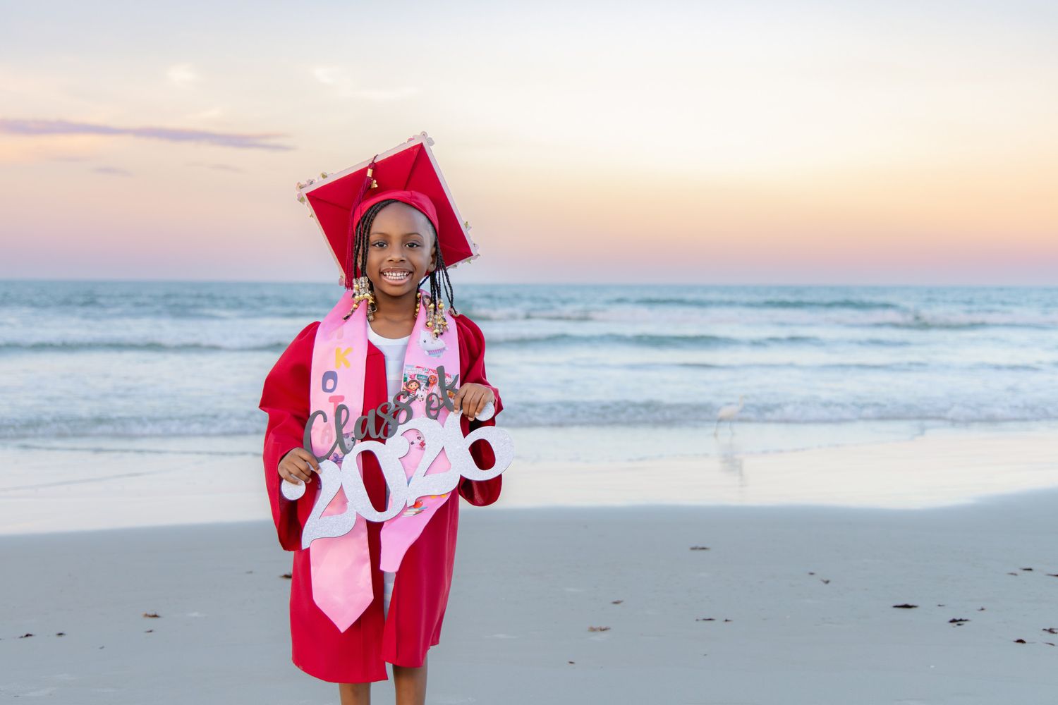 Young girl in red cap and gown holds graduation sign on sandy beach at dusk.