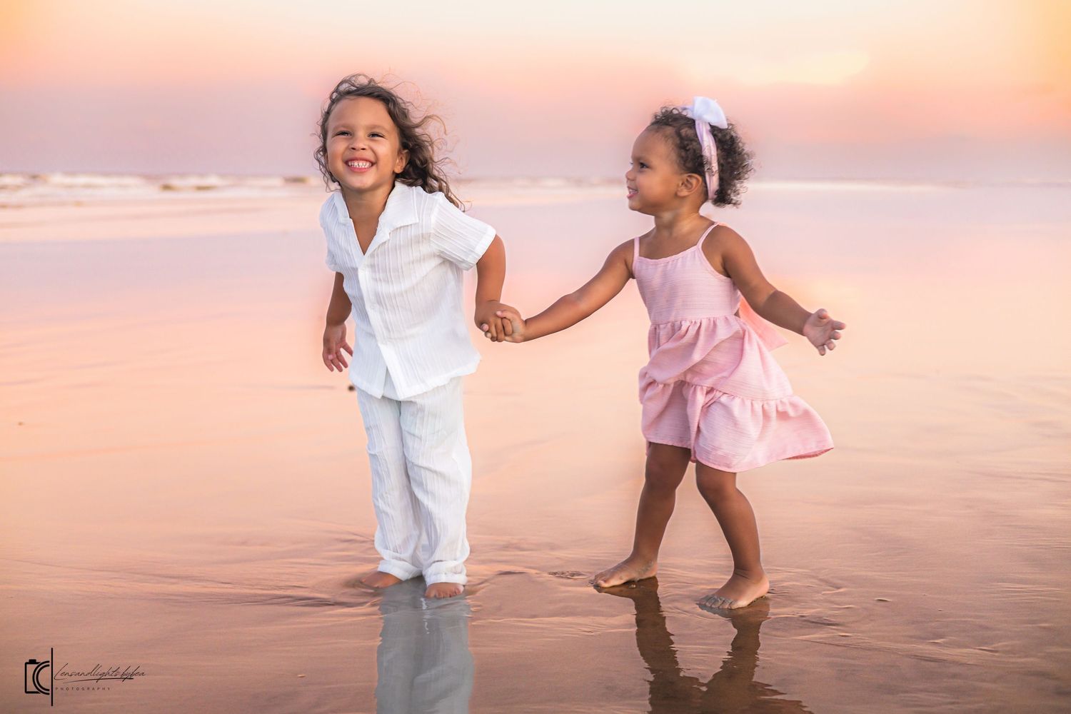 Children in white and pink outfits playing and running together on a beach at sunset with warm orange sky.