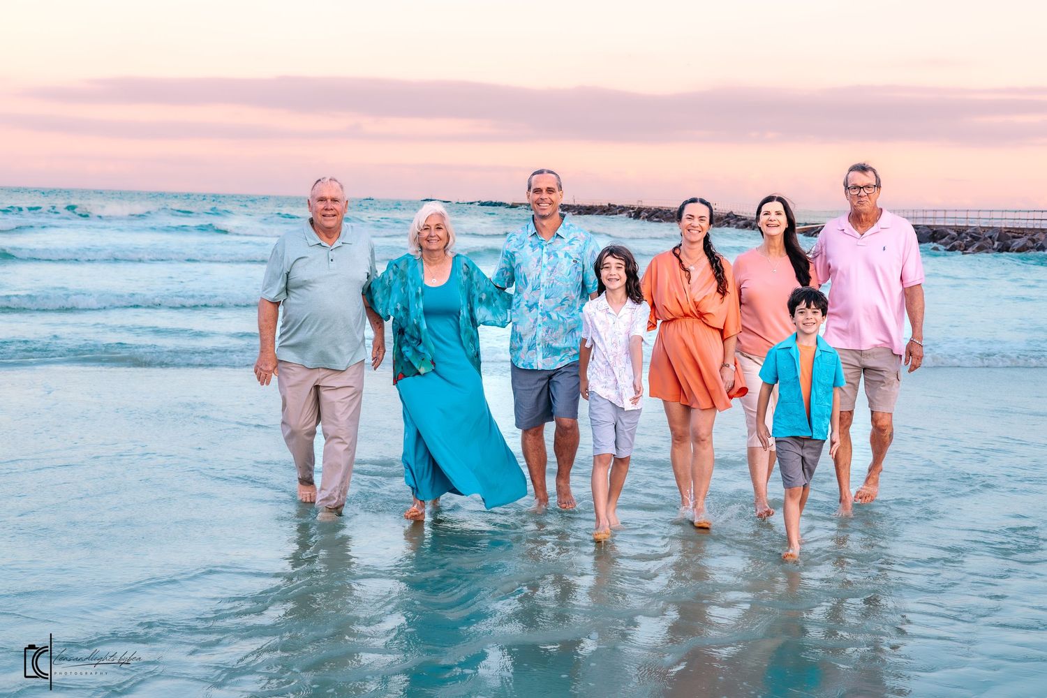 A group of people standing together in the shallow ocean water at sunset with pink and blue sky in the background.