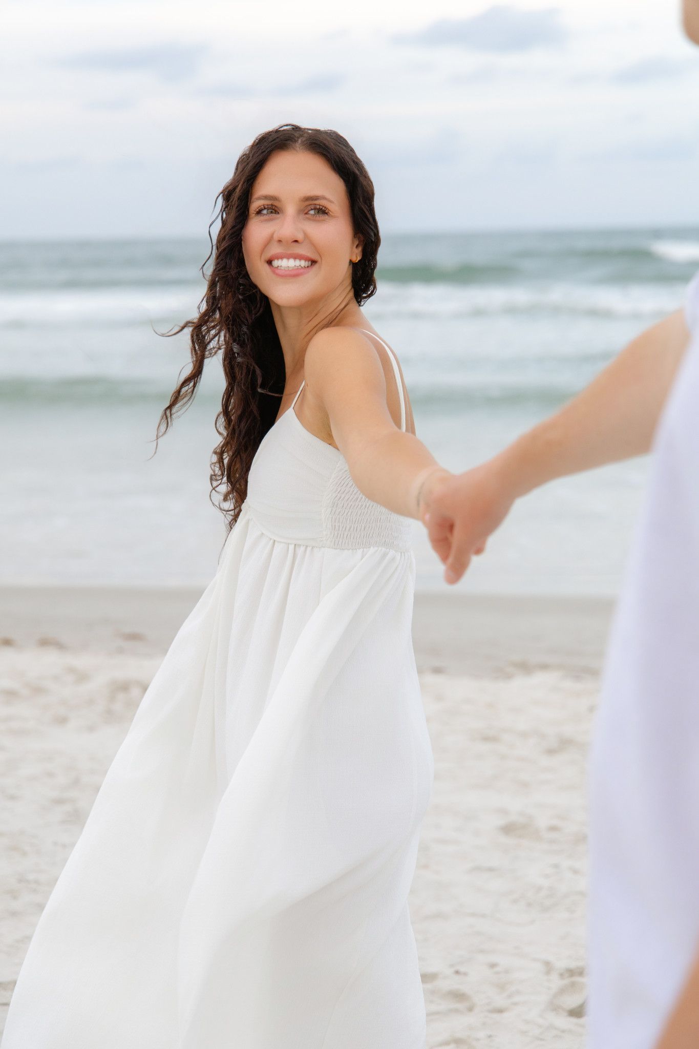Smiling brunette woman in white dress holding hands with partner on sandy beach with ocean waves.