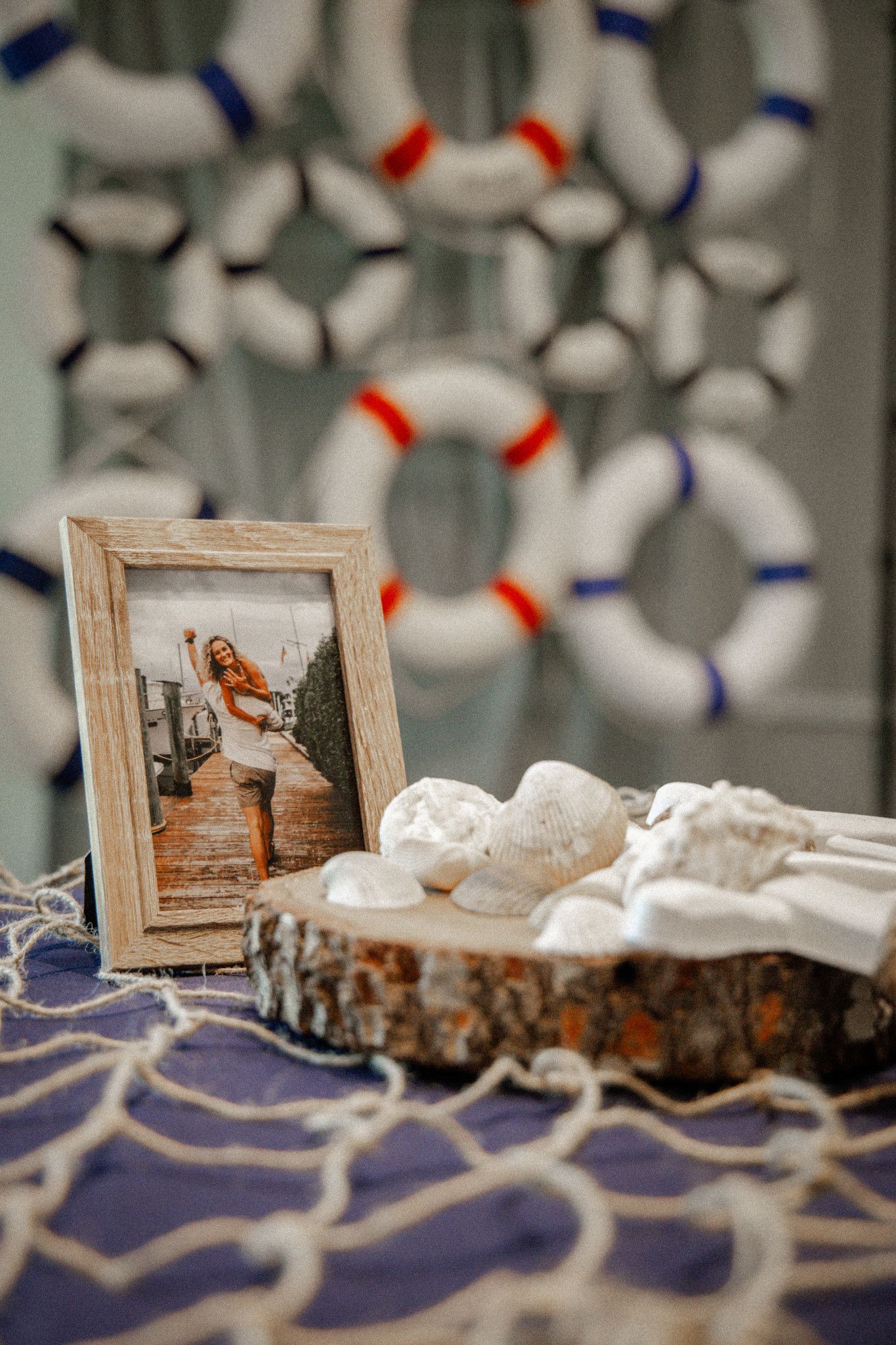 Nautical wedding decor with framed photo, seashells on wood slice, fish net, and life preservers in background.