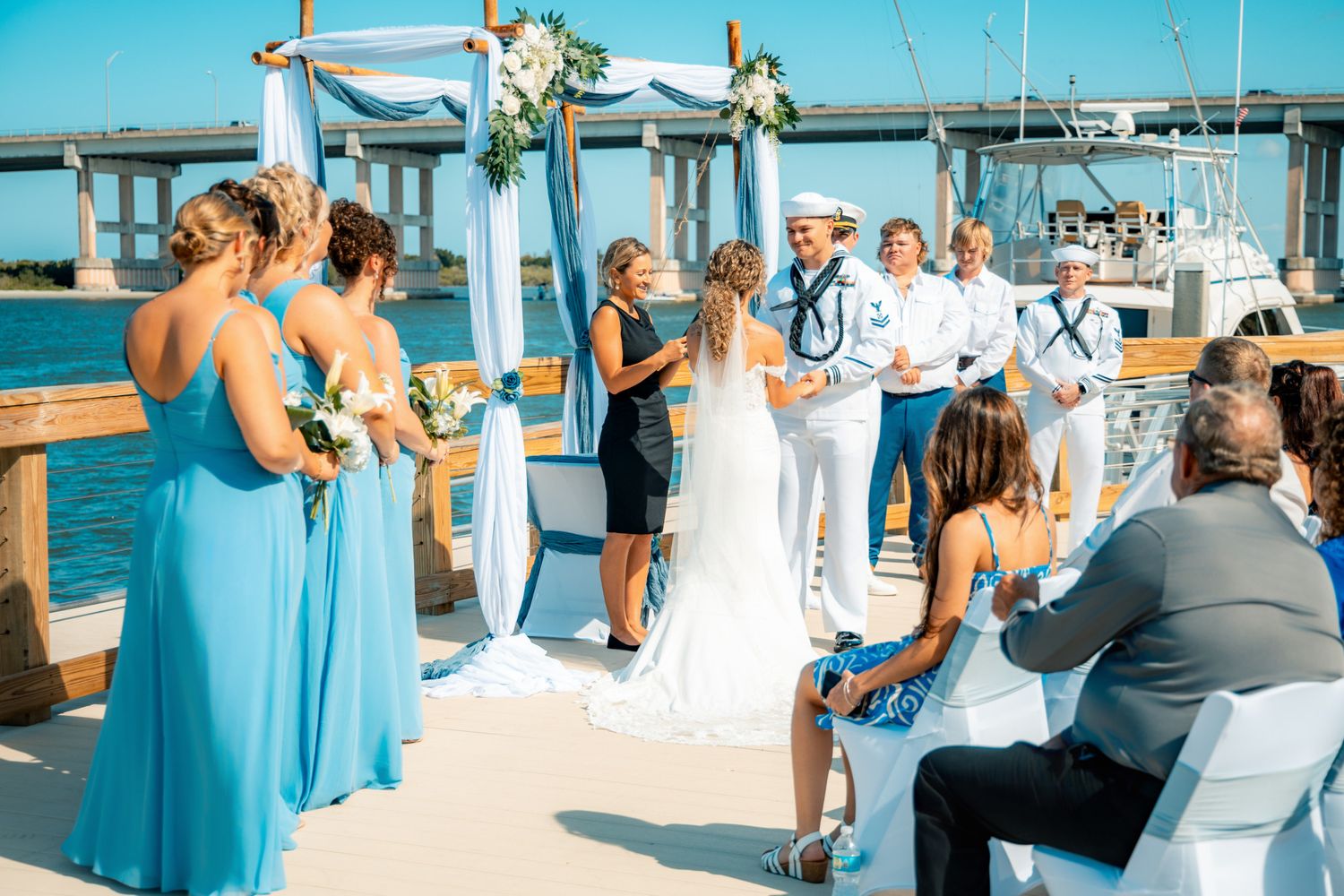 Outdoor waterfront wedding ceremony on a dock with bridesmaids in blue dresses and groom in sailor uniform.