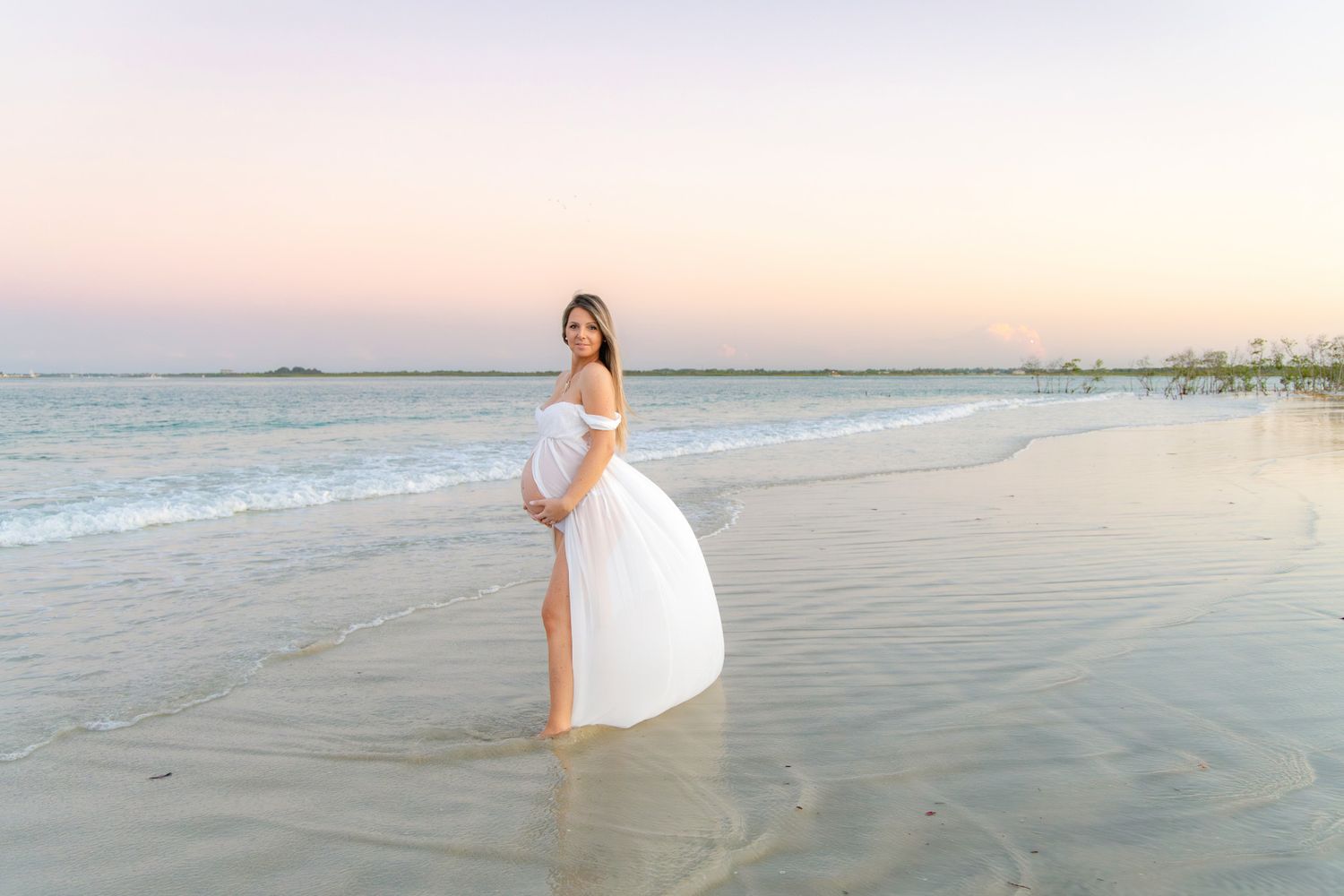Pregnant woman in white dress standing on beach at sunset with ocean waves.