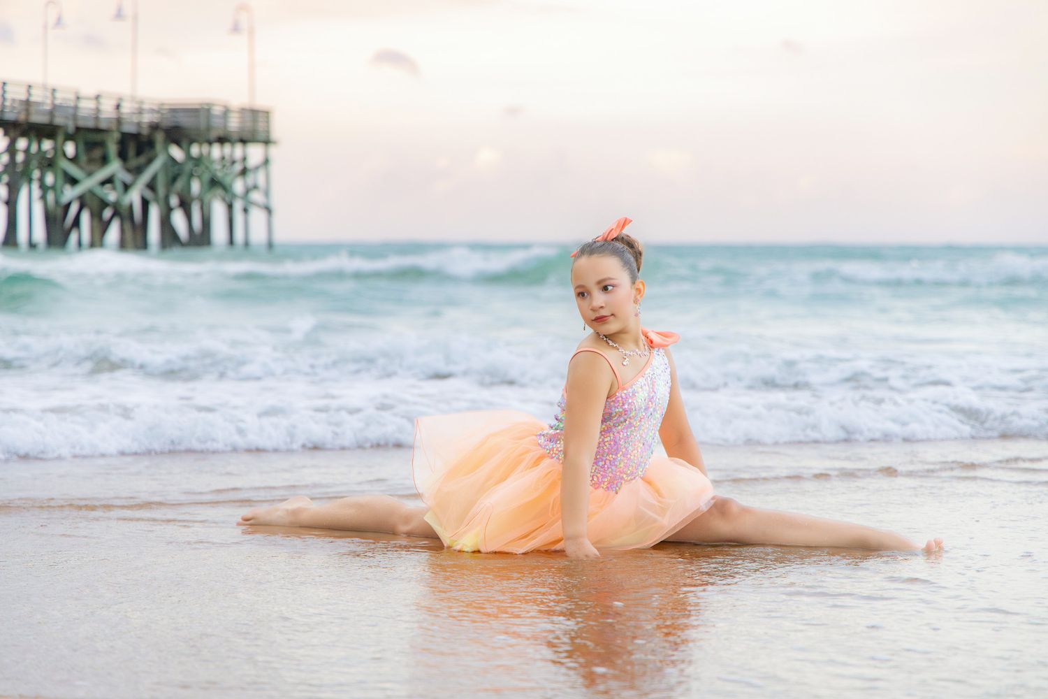 Young ballerina in pink tutu performs splits on sandy beach near pier at sunset.
