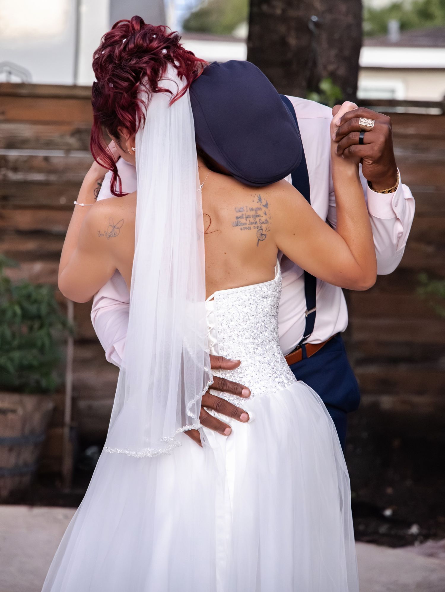 A couple in wedding attire embraces outdoors, with the bride wearing a white beaded gown and burgundy-colored hair.