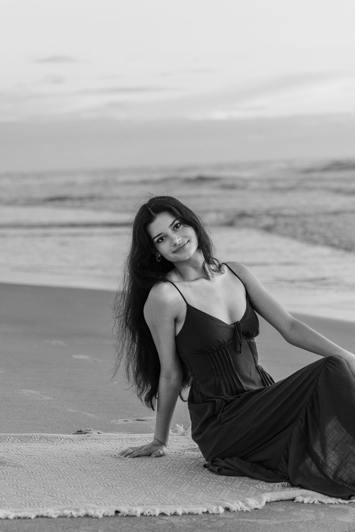 Black and white photo of a young woman in a dark dress sitting on a beach with ocean waves in the background.