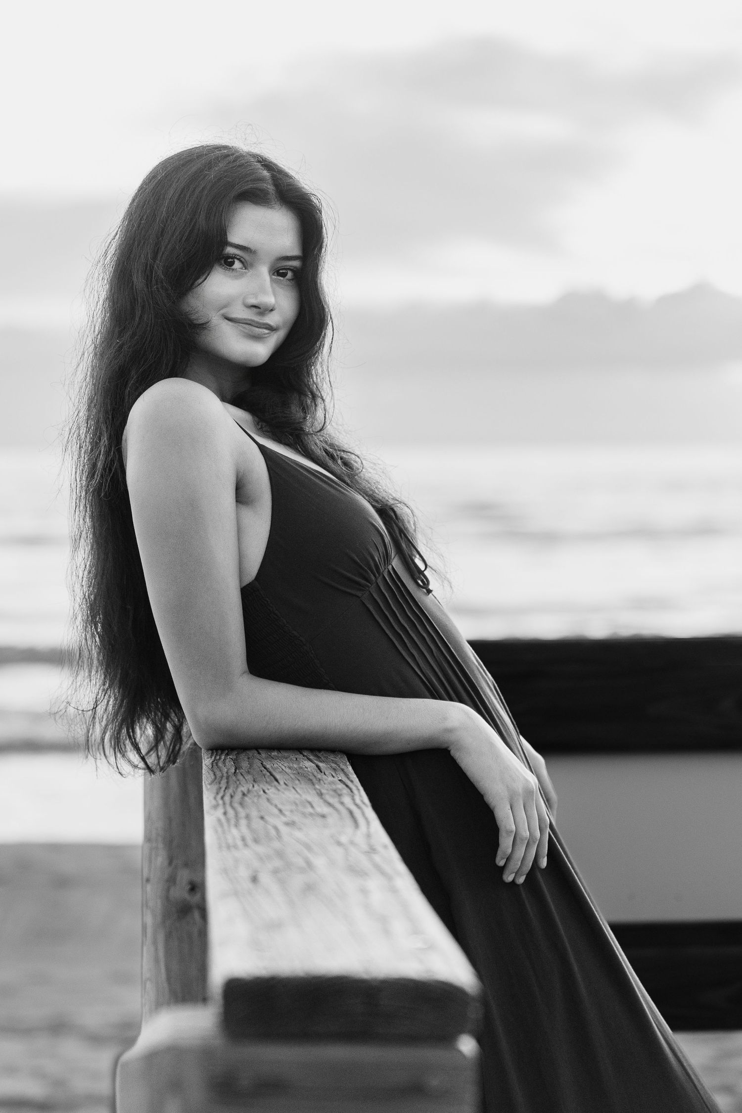 Young woman with long dark hair in a flowing dress leans on a wooden railing by the ocean in black and white.