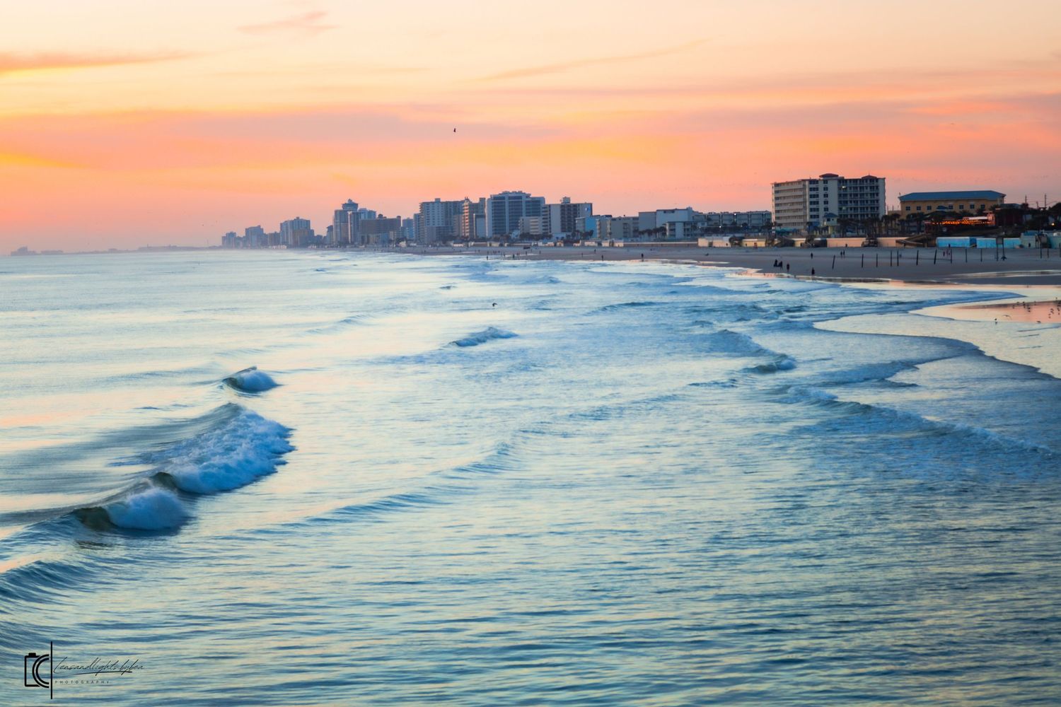 Waves roll onto a sandy beach at sunset with a coastal cityscape silhouetted against an orange sky.