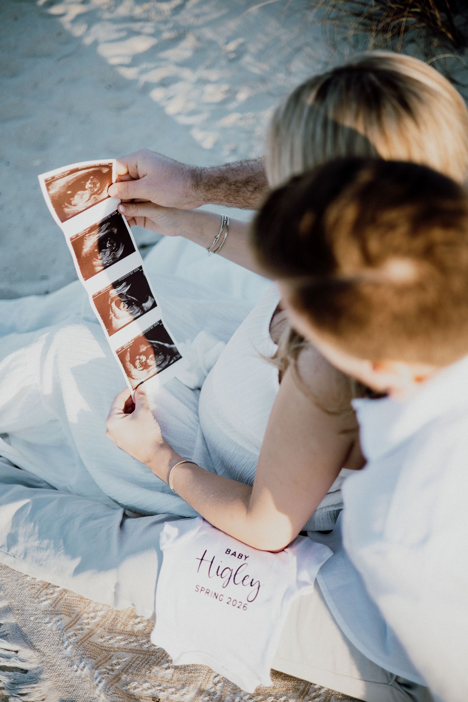 Someone looks at ultrasound photos while sitting on light blue bedding during a pregnancy announcement photoshoot.
