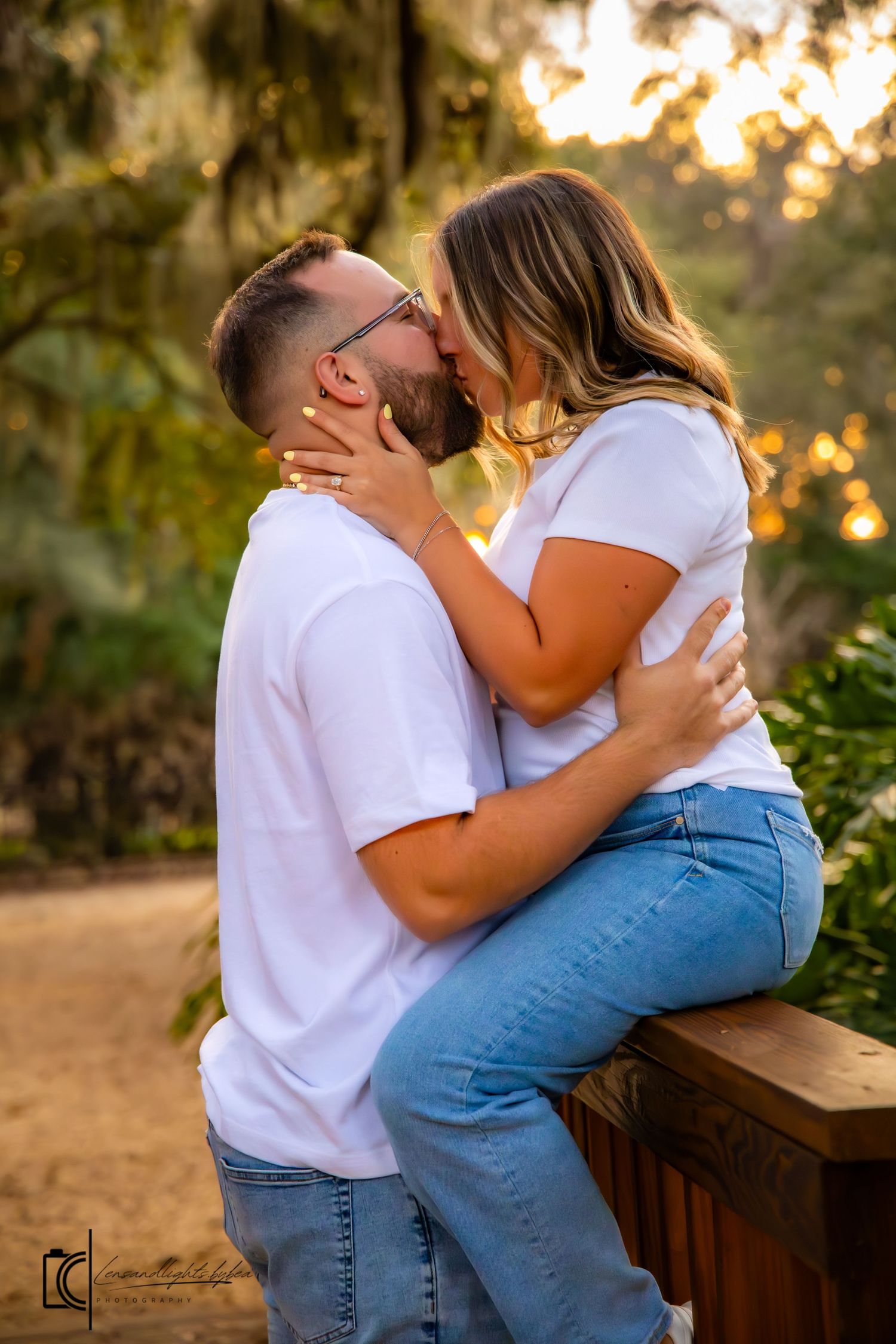 A romantic couple in white and blue outfits share an intimate moment on a wooden bench at sunset.