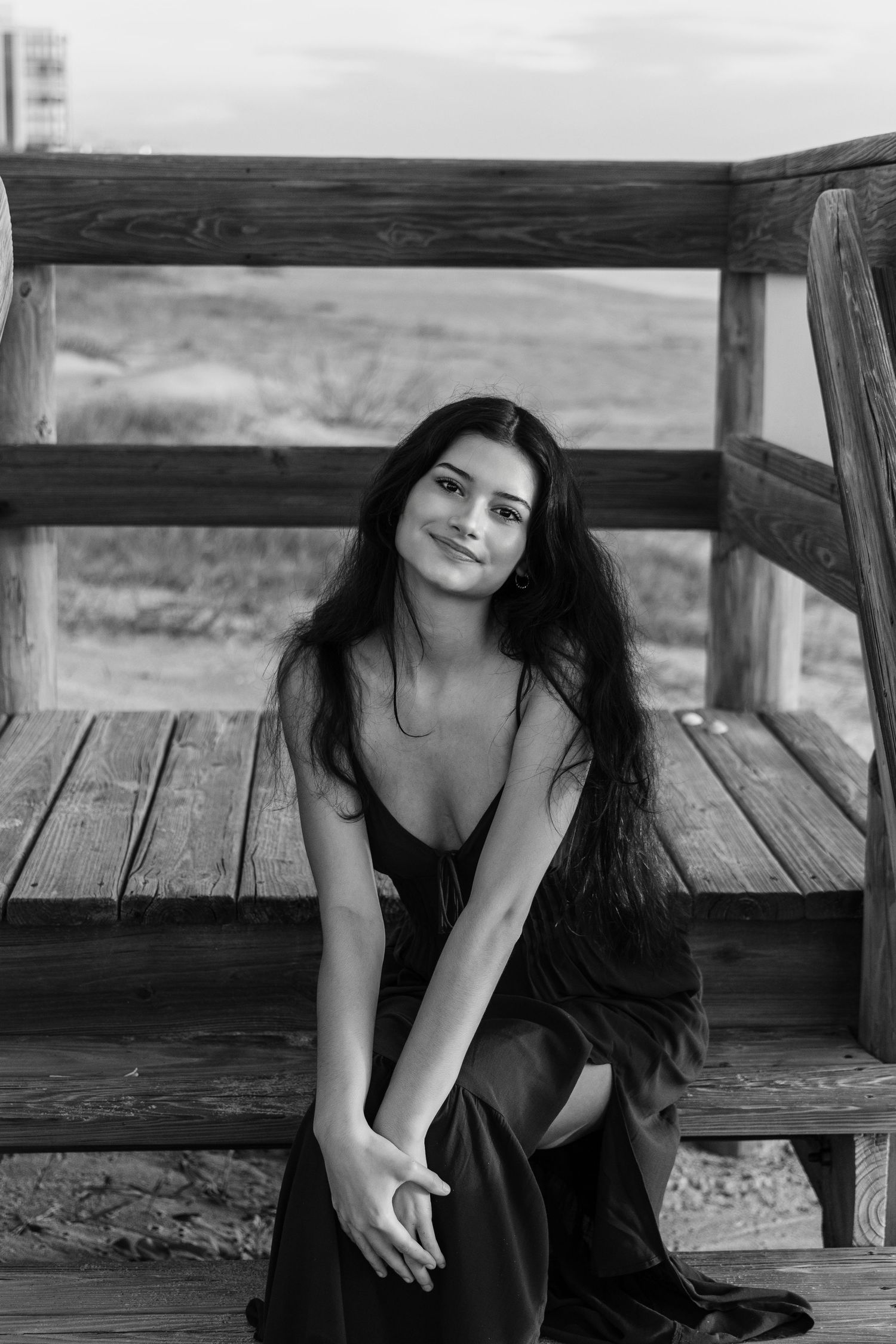 Black and white photo of a young woman in a dark dress sitting on wooden beach steps, smiling softly at camera.