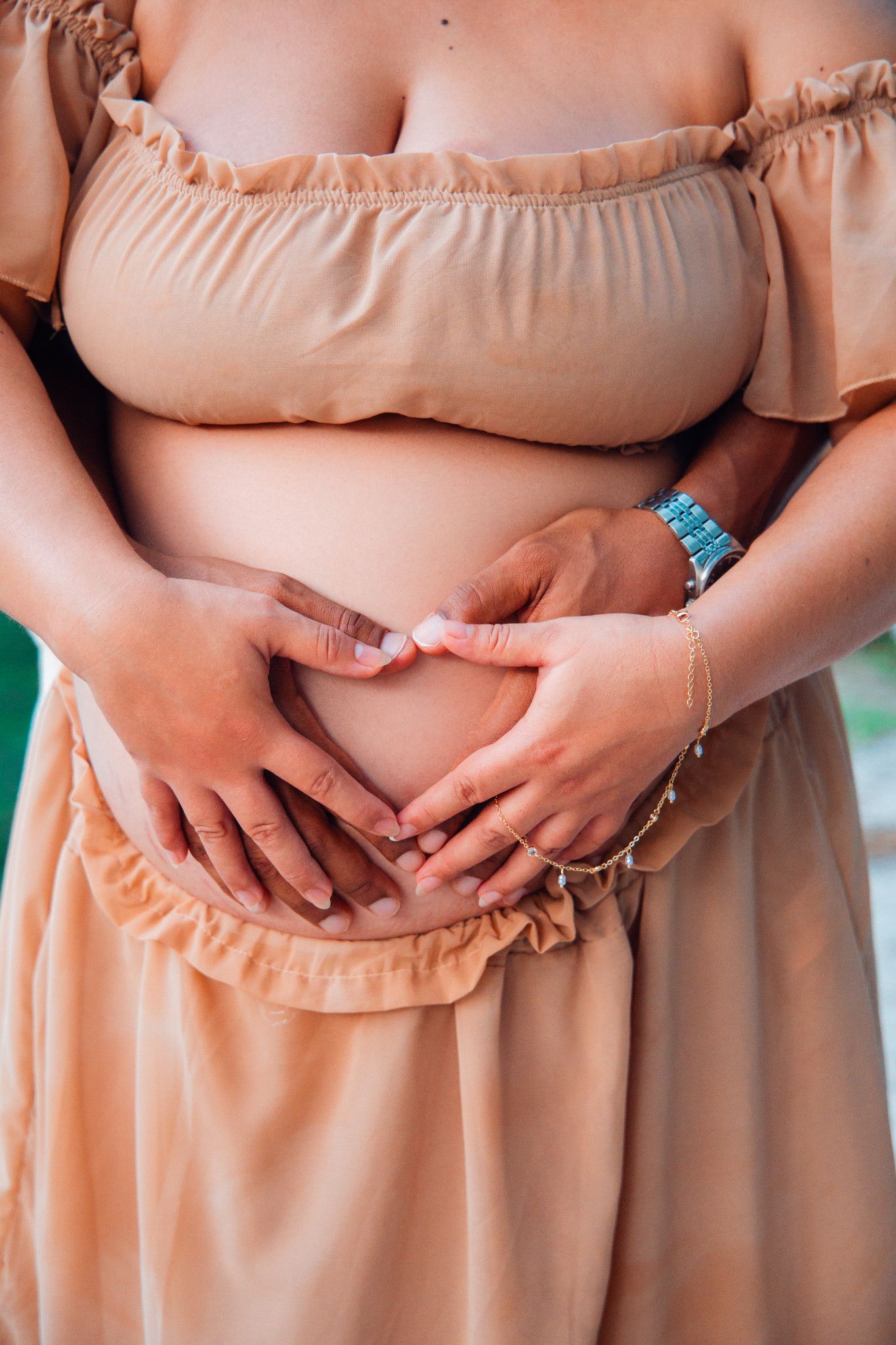 Close-up of couple forming heart shape with hands on pregnant belly in peach off-shoulder maternity dress.