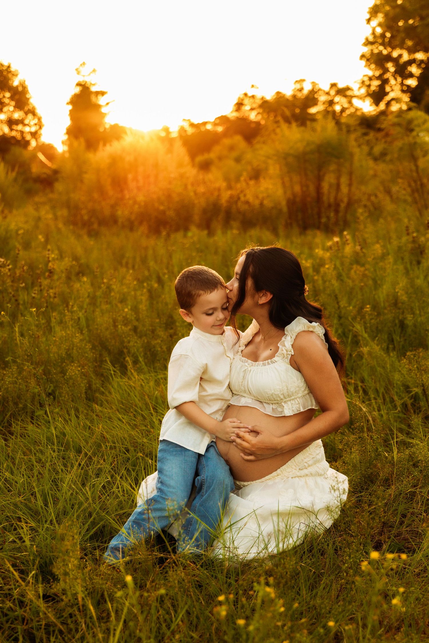 Pregnant mother sits in golden sunset field while young son kisses her cheek, both wearing white, hands on baby bump.