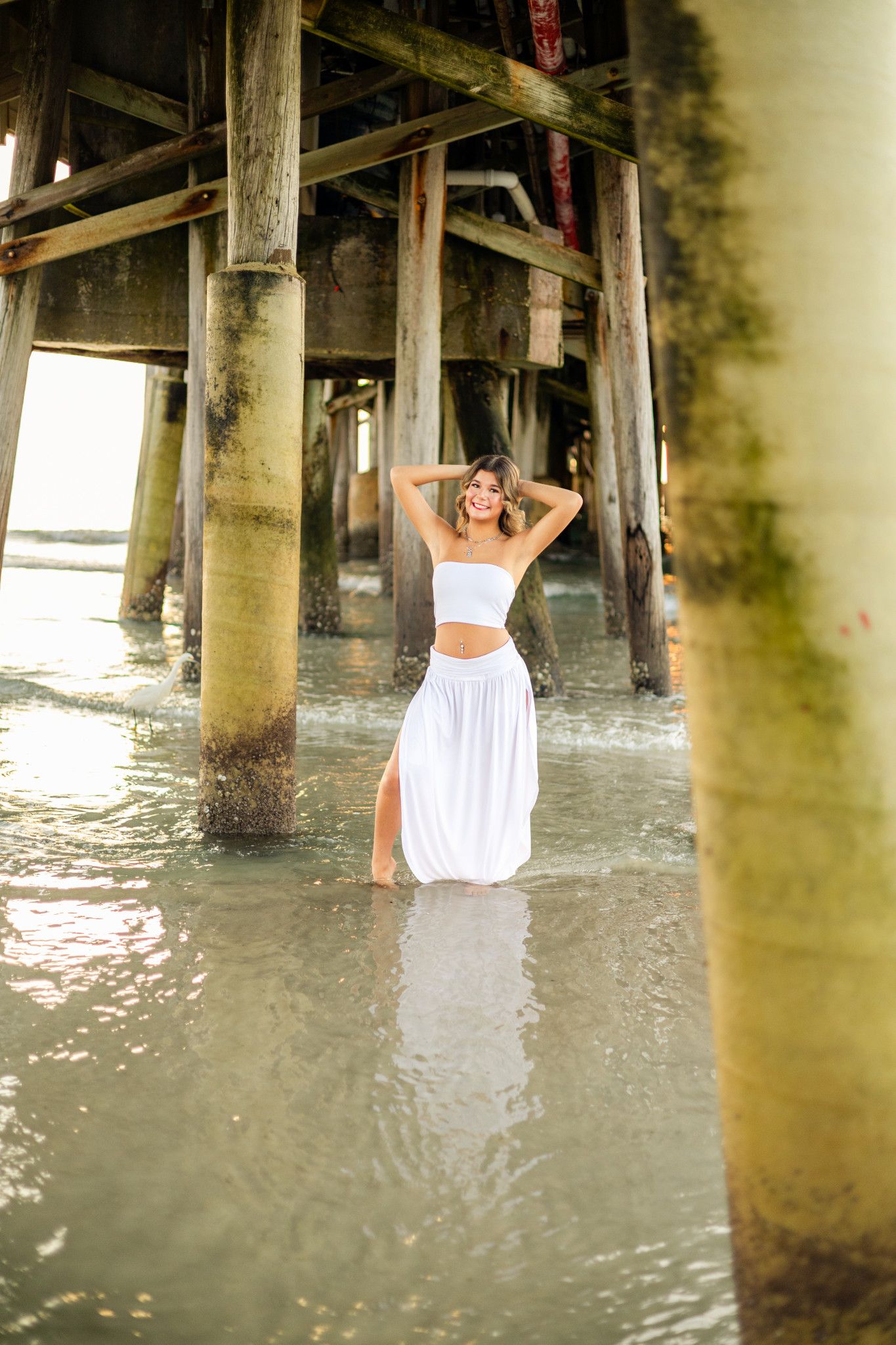 Woman in white crop top and maxi skirt standing in shallow water beneath a wooden pier.