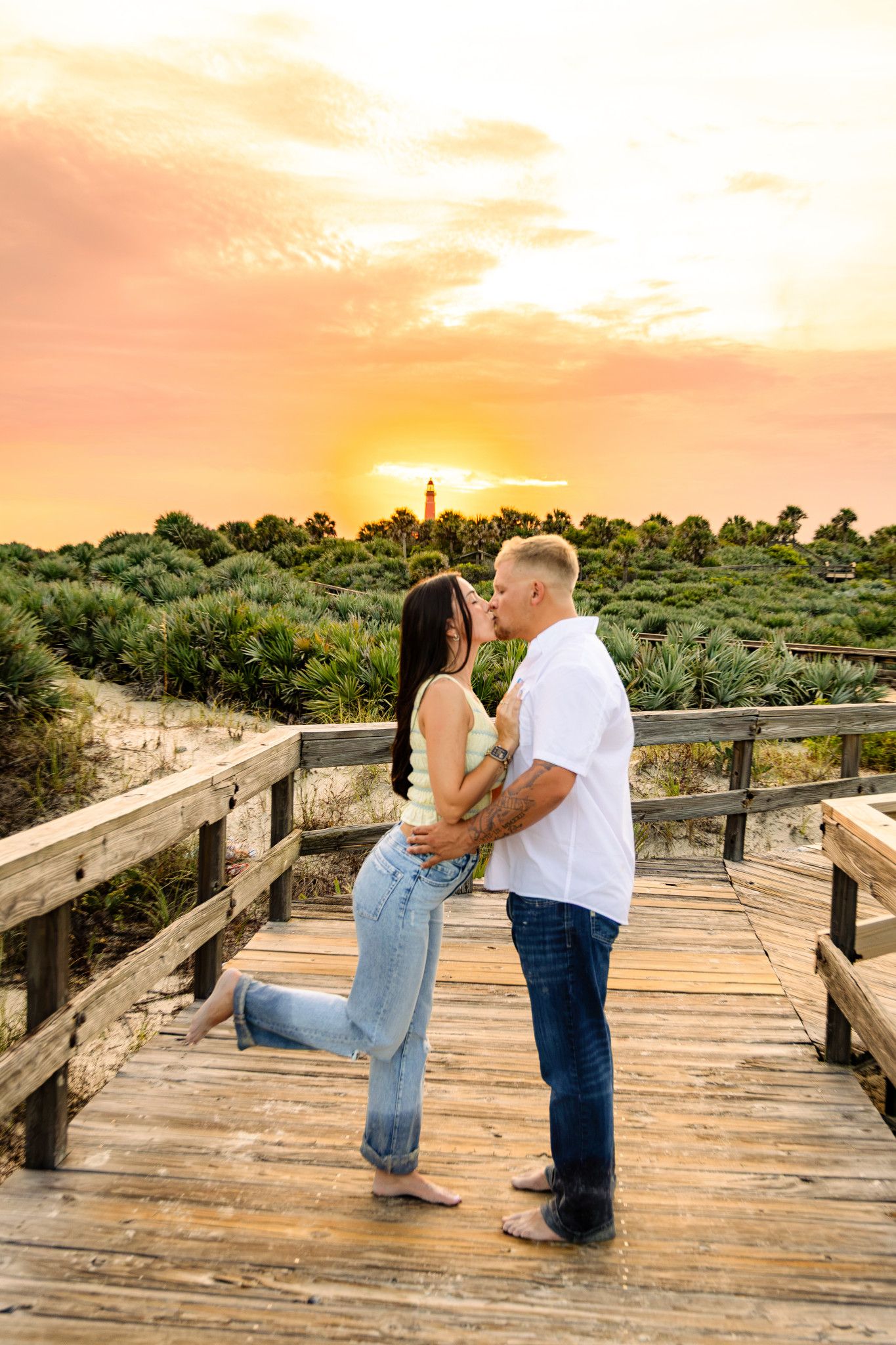 Couple sharing a romantic kiss on a wooden boardwalk at sunset with a lighthouse in the background.