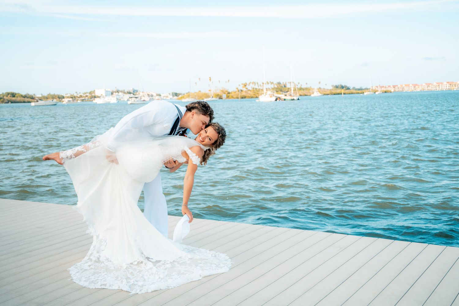 Groom dips bride in white gown on waterfront dock during romantic outdoor wedding photo session.