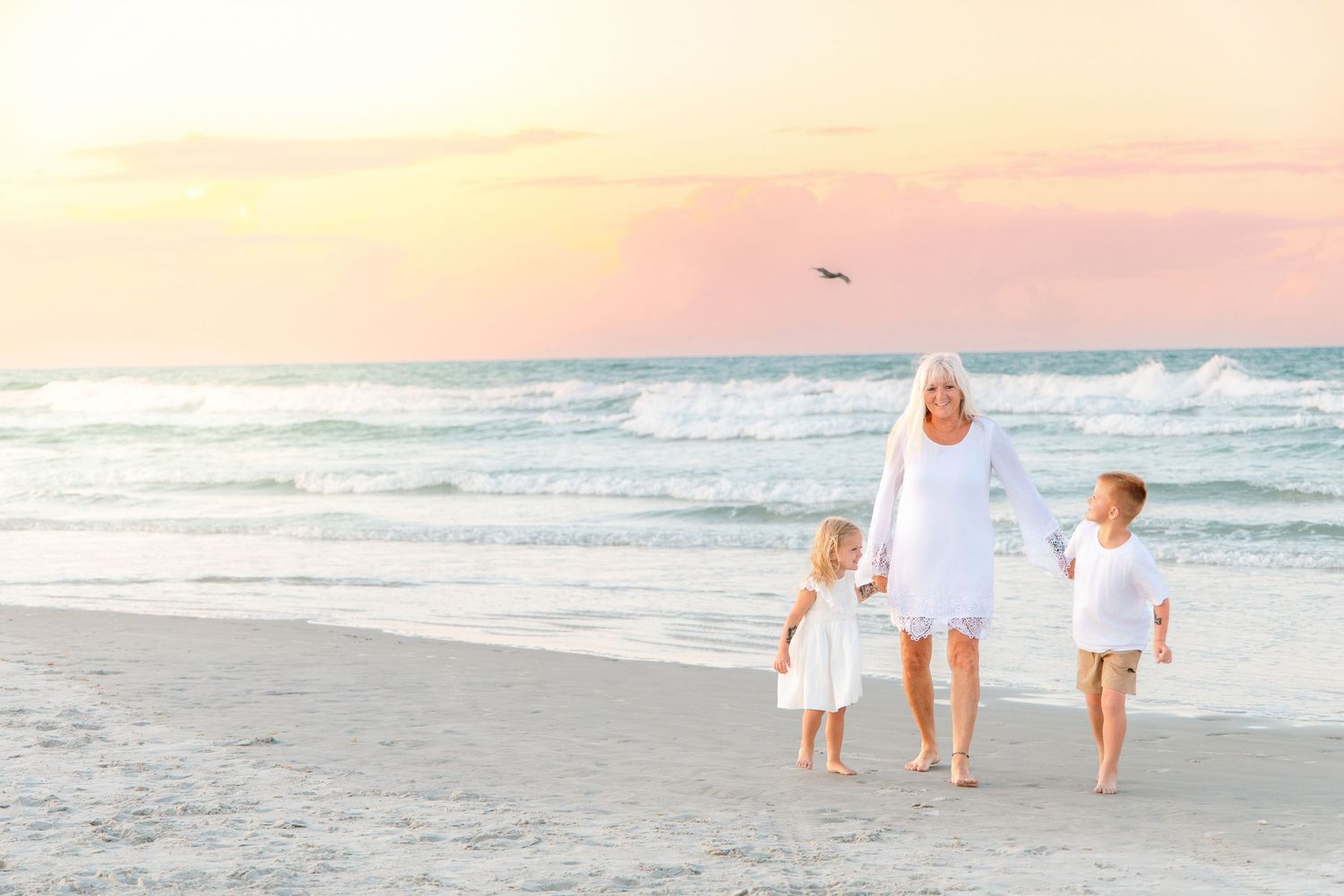 Grandmother walks on a sandy beach at sunset with two young children in white outfits, ocean waves in the background.