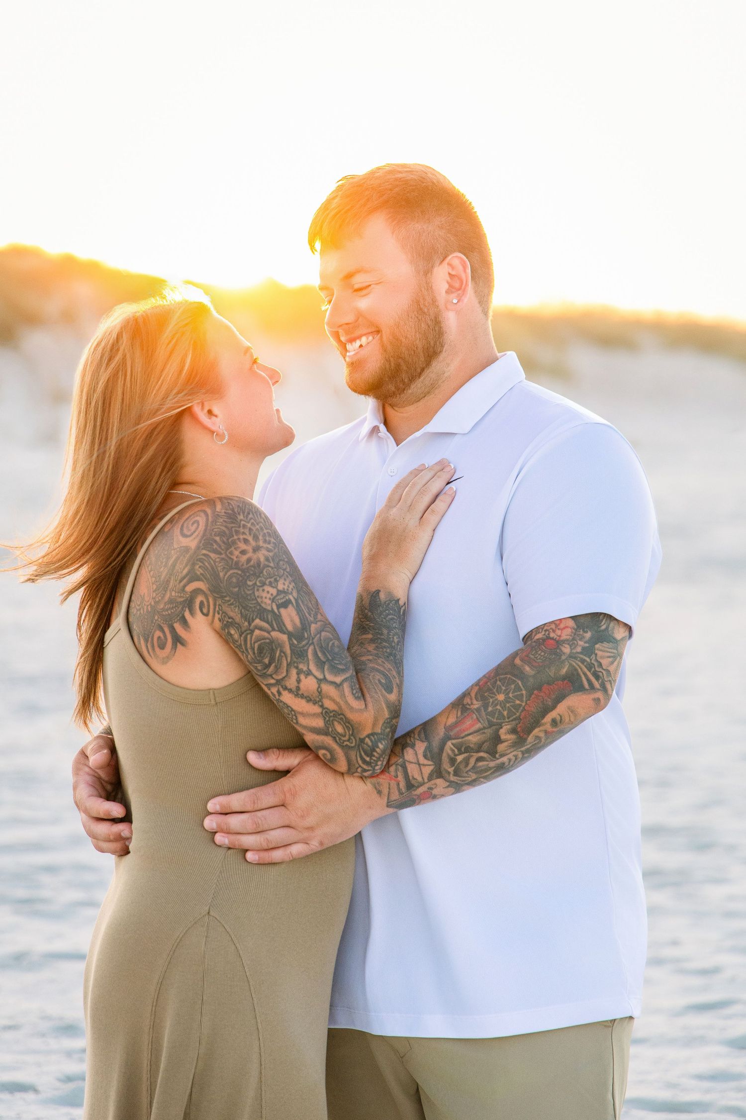 Tattooed couple embraces and smiles at each other on a beach during golden hour sunset.