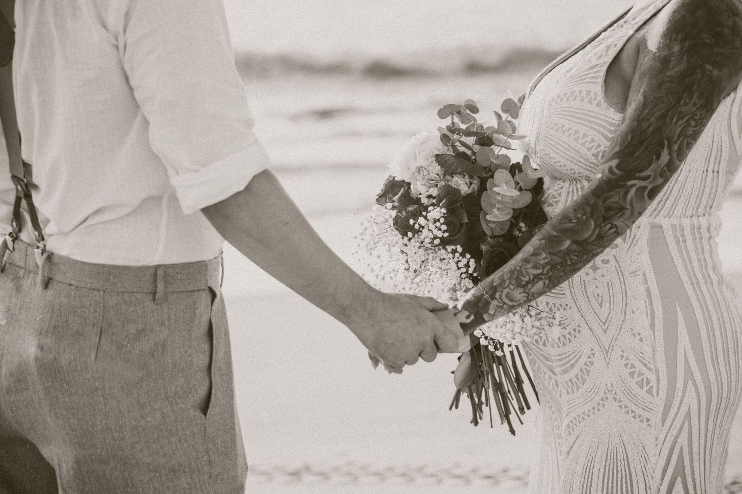 Black and white close-up of bride and groom holding a floral bouquet together on the beach.
