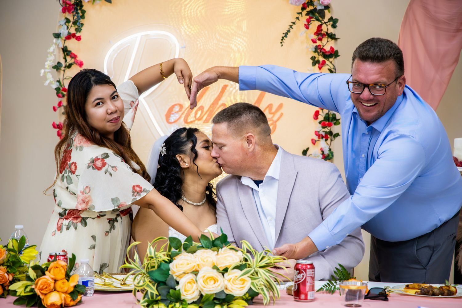 Couple sharing a kiss at wedding reception table with floral decor while guests celebrate behind them.