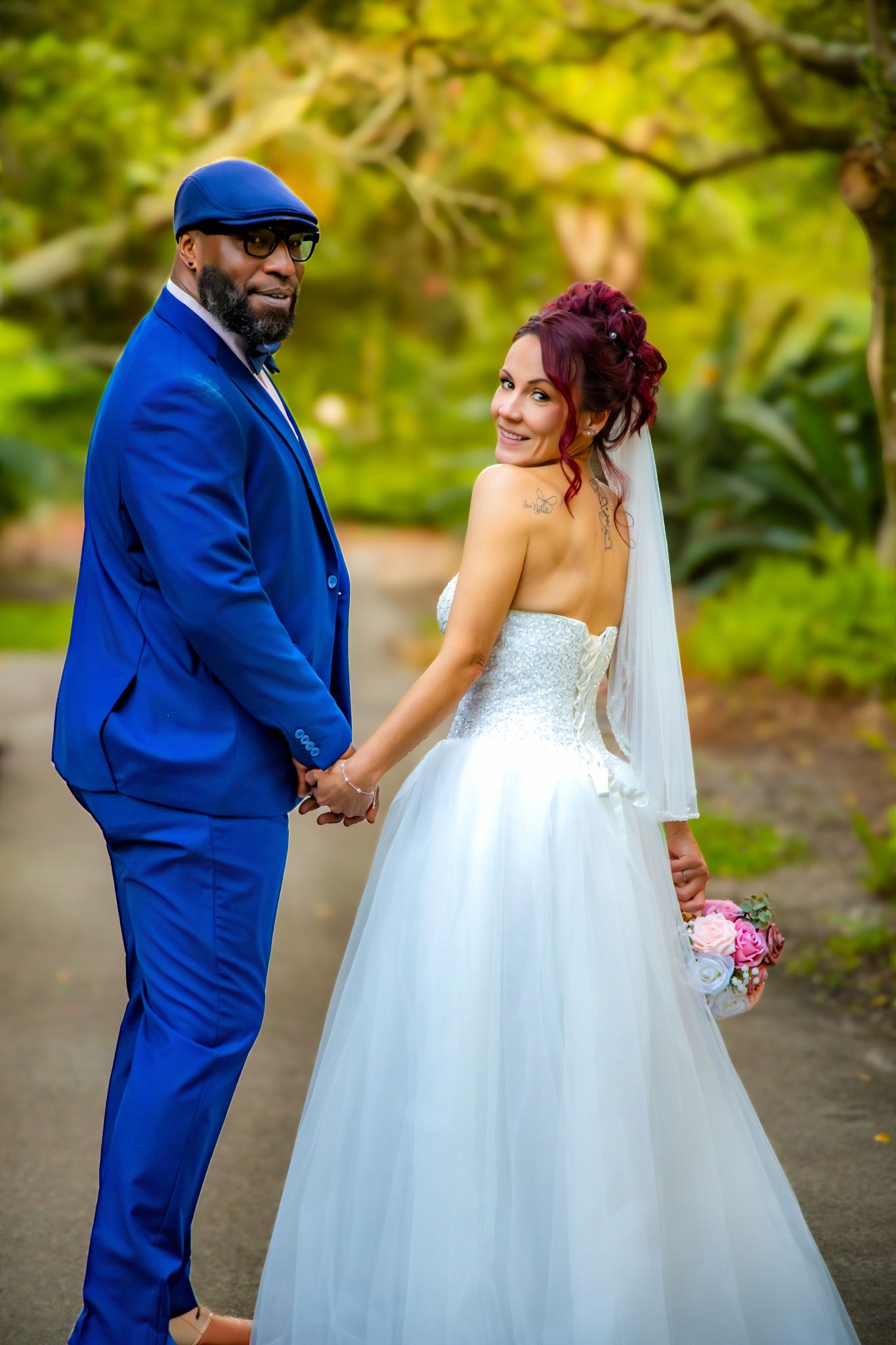 A bride in a white gown and groom in royal blue suit share tender moments during outdoor wedding photos in a garden setting.